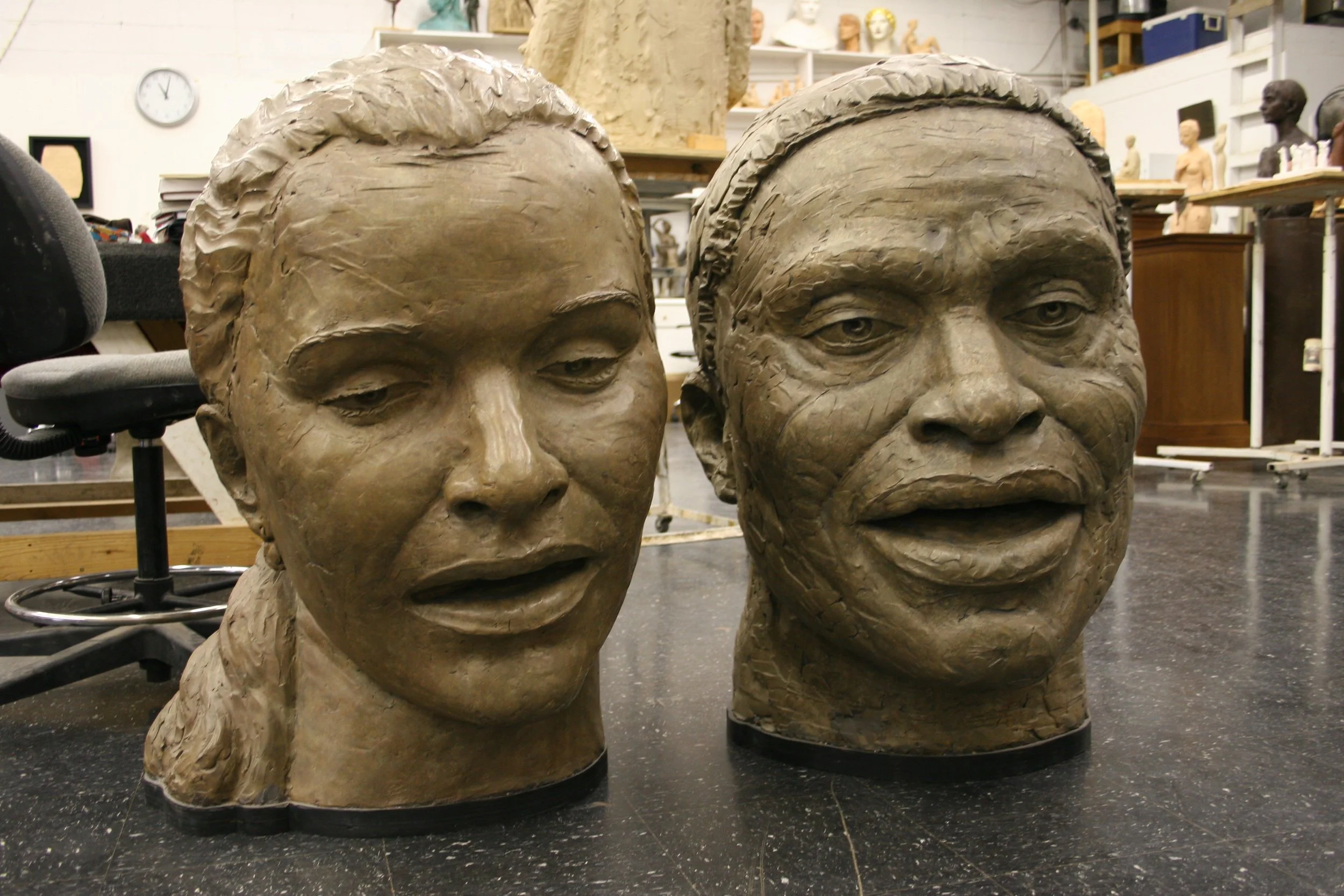 Bronze busts of a women and man with detailed features and textured hair, in an artist's studio with shelves containing sculptures and busts in the background.  Cultural Heroes