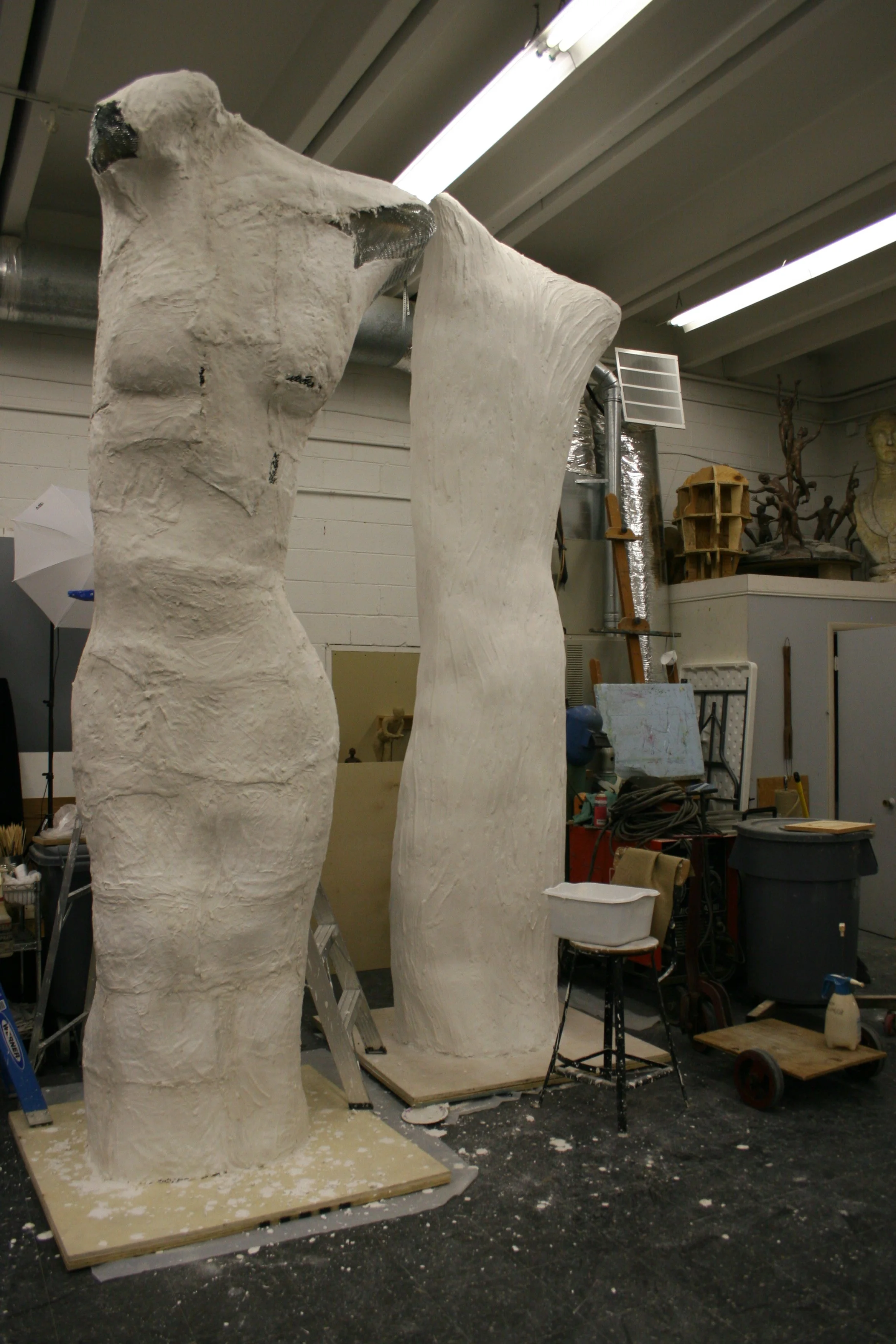 Two large, unfinished sculptures of human torsos in an artist's workshop, surrounded by tools, materials, and other sculptures.