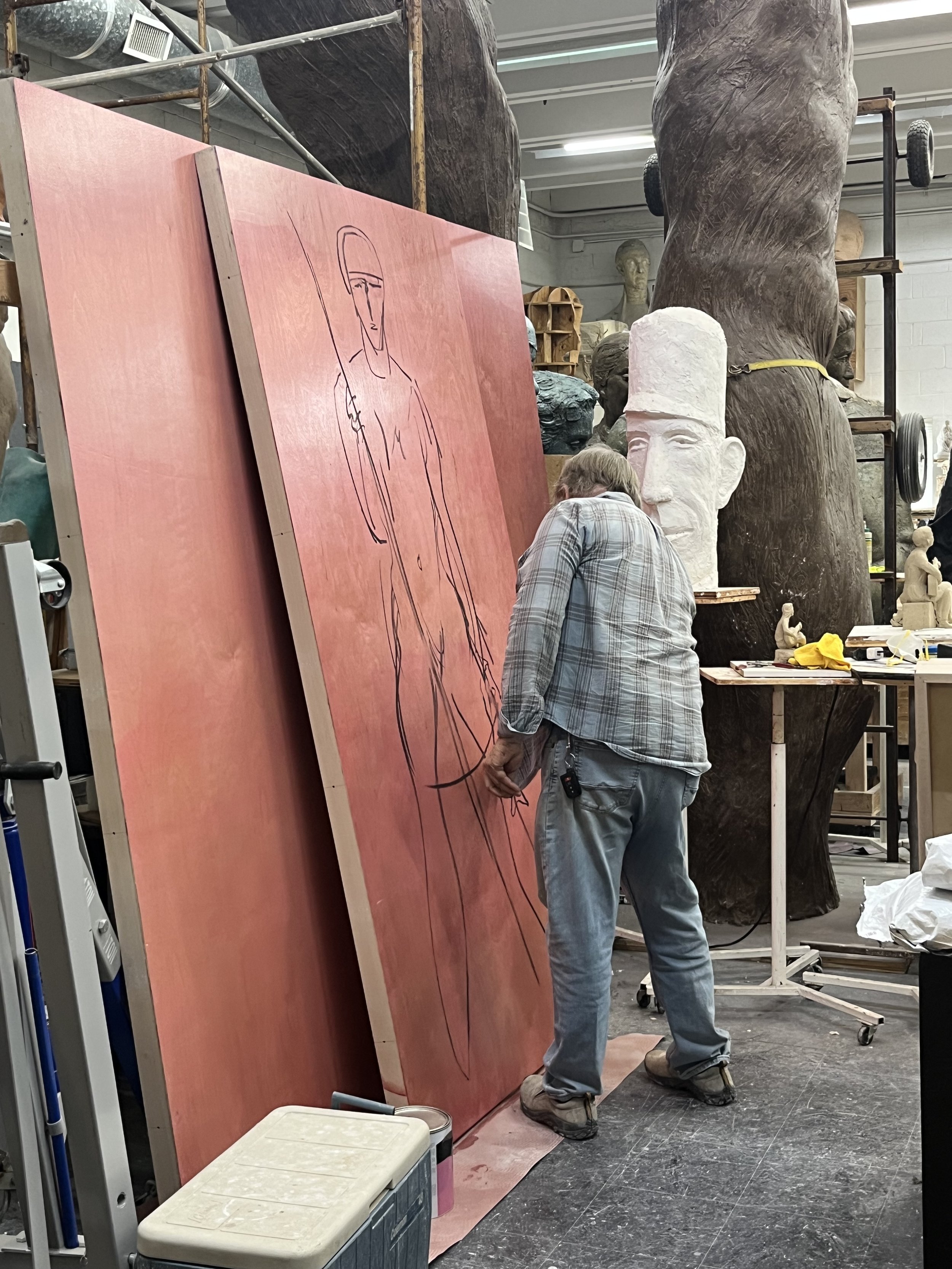 An artist in a studio working on a large pink canvas with a simple line drawing of a person. Various sculptures and busts are visible in the background, along with art supplies and equipment.