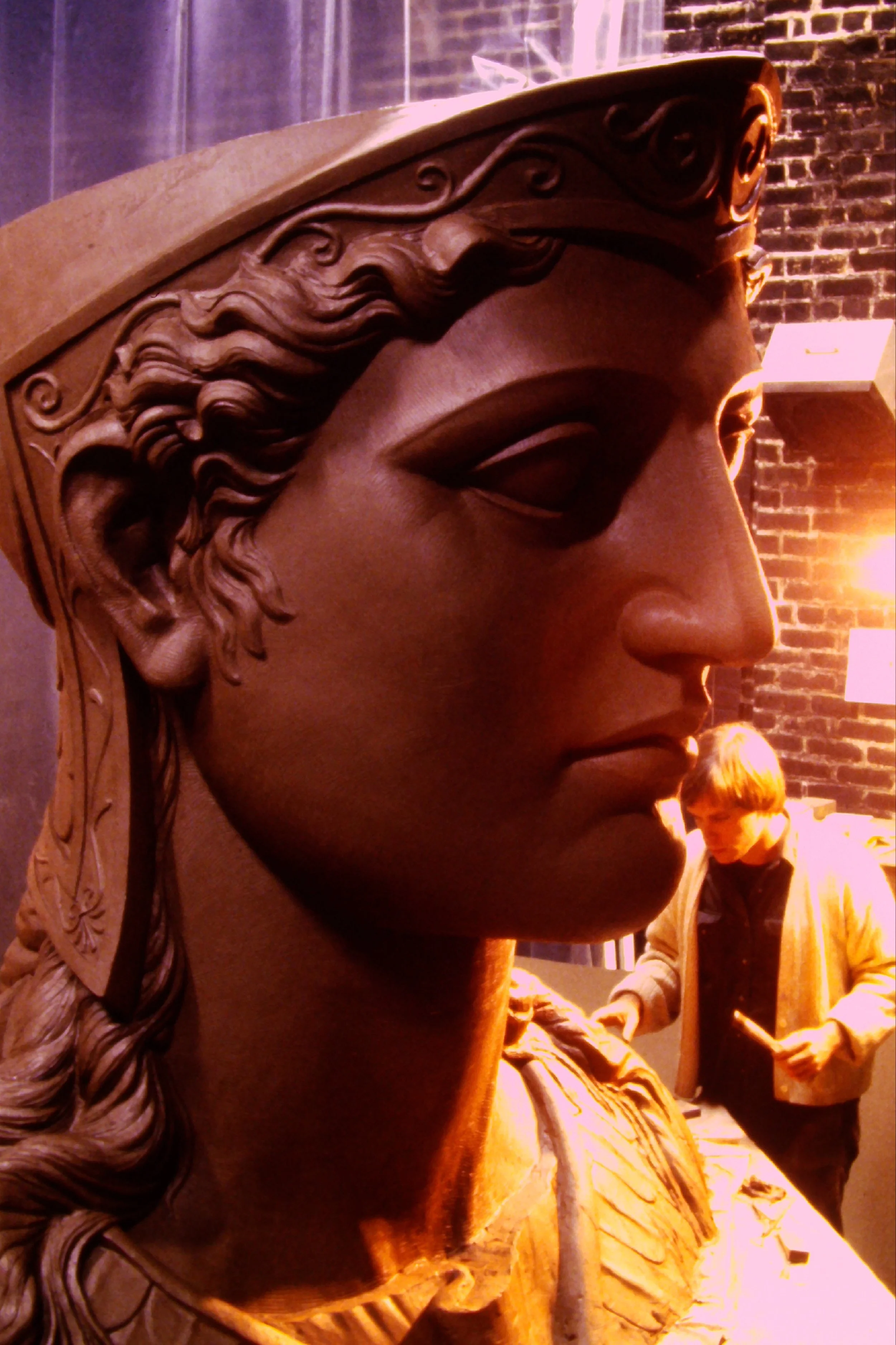 Close-up of a large, detailed sculpture of a face, featuring a prominent brow, eye, nose, and curly hair, with a brick wall and people in the background. Athena Parthenos in Parthenon Museum Nashville. 