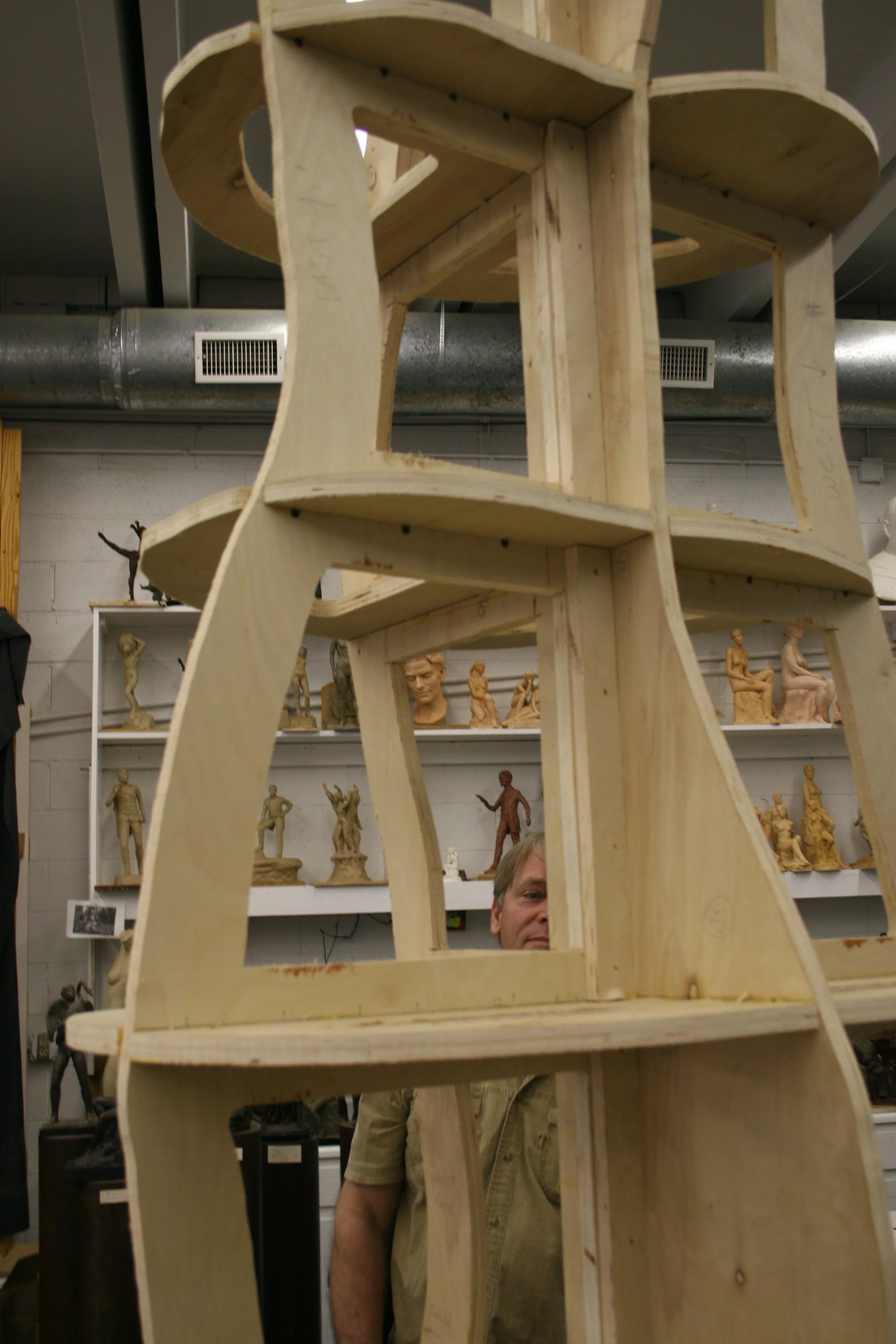A wooden sculpture frame being built in a workshop, with shelves displaying various small sculptures and figurines in the background.