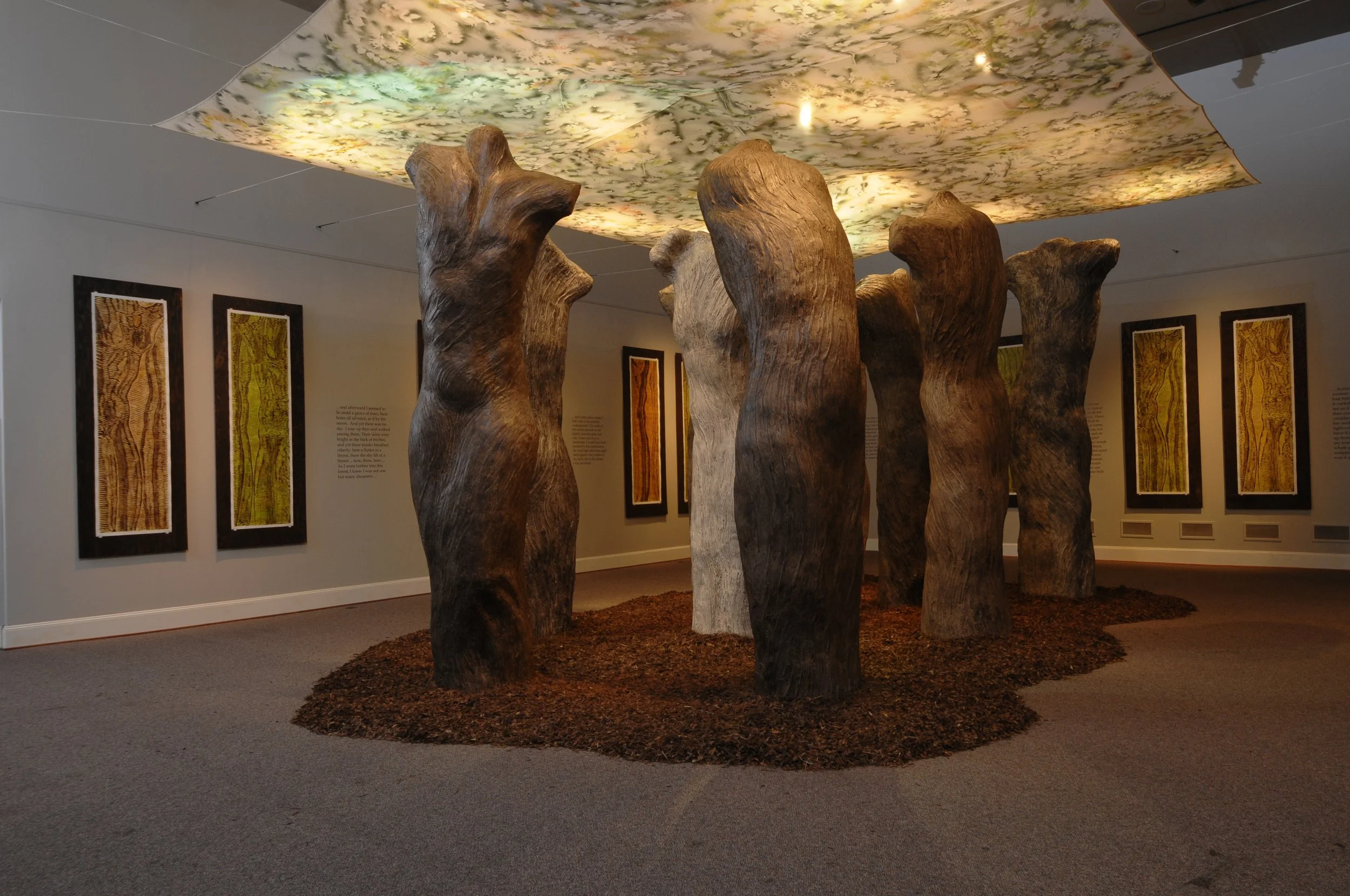 An art installation featuring large, carved wooden poles arranged in a circle on a dirt base, with framed art pieces on the walls behind it, and a illuminated floral patterned ceiling overhead. Dream Forest installation 