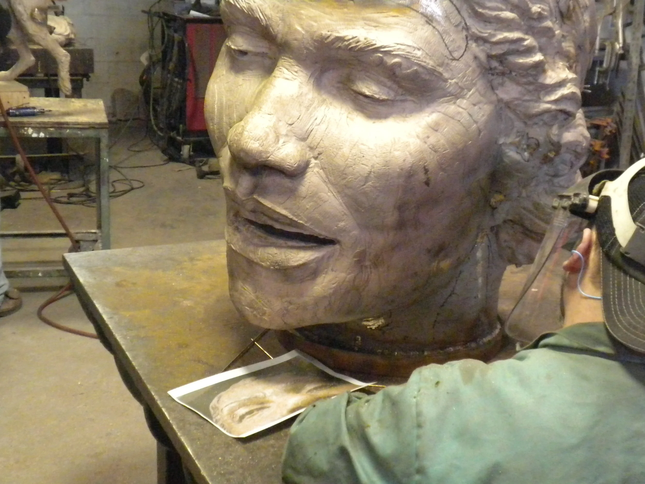 A large bronze sculpture of a human face in progress, placed on a worktable in a foundry with various tools and equipment in the background.