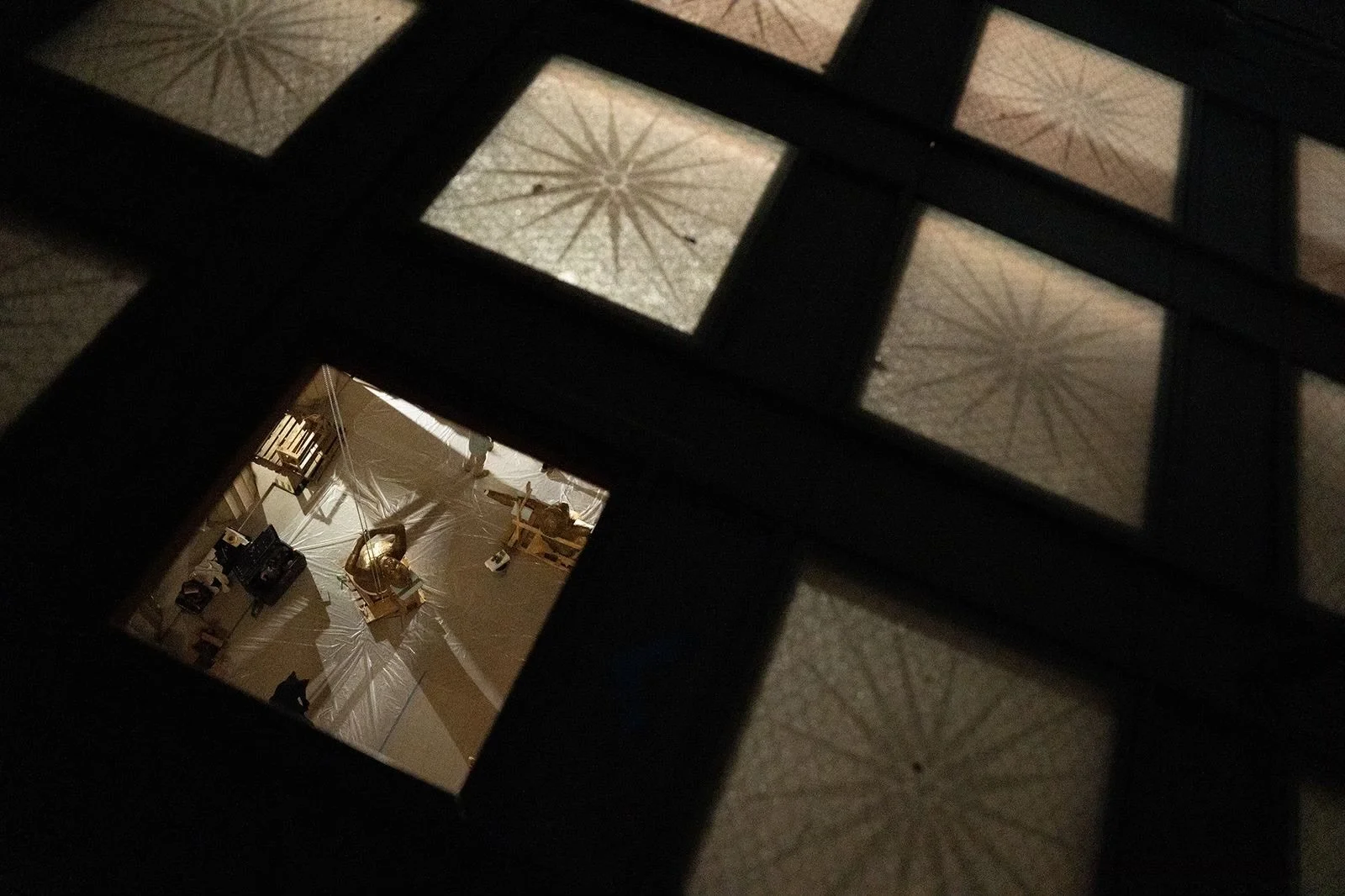 View through a glass window showing a room with furniture, a person, and various objects, framed by a dark grid of squares and rectangles. Parthenon Museum. 