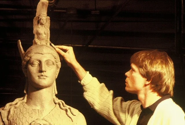 A person sculpting a large clay statue of a woman's head with a headdress.