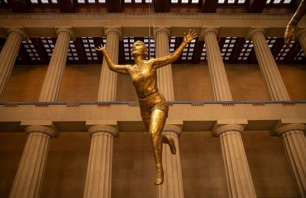 Gold statue of a woman in mid-jump with arms raised, located inside a building with tall, fluted columns and a coffered ceiling.