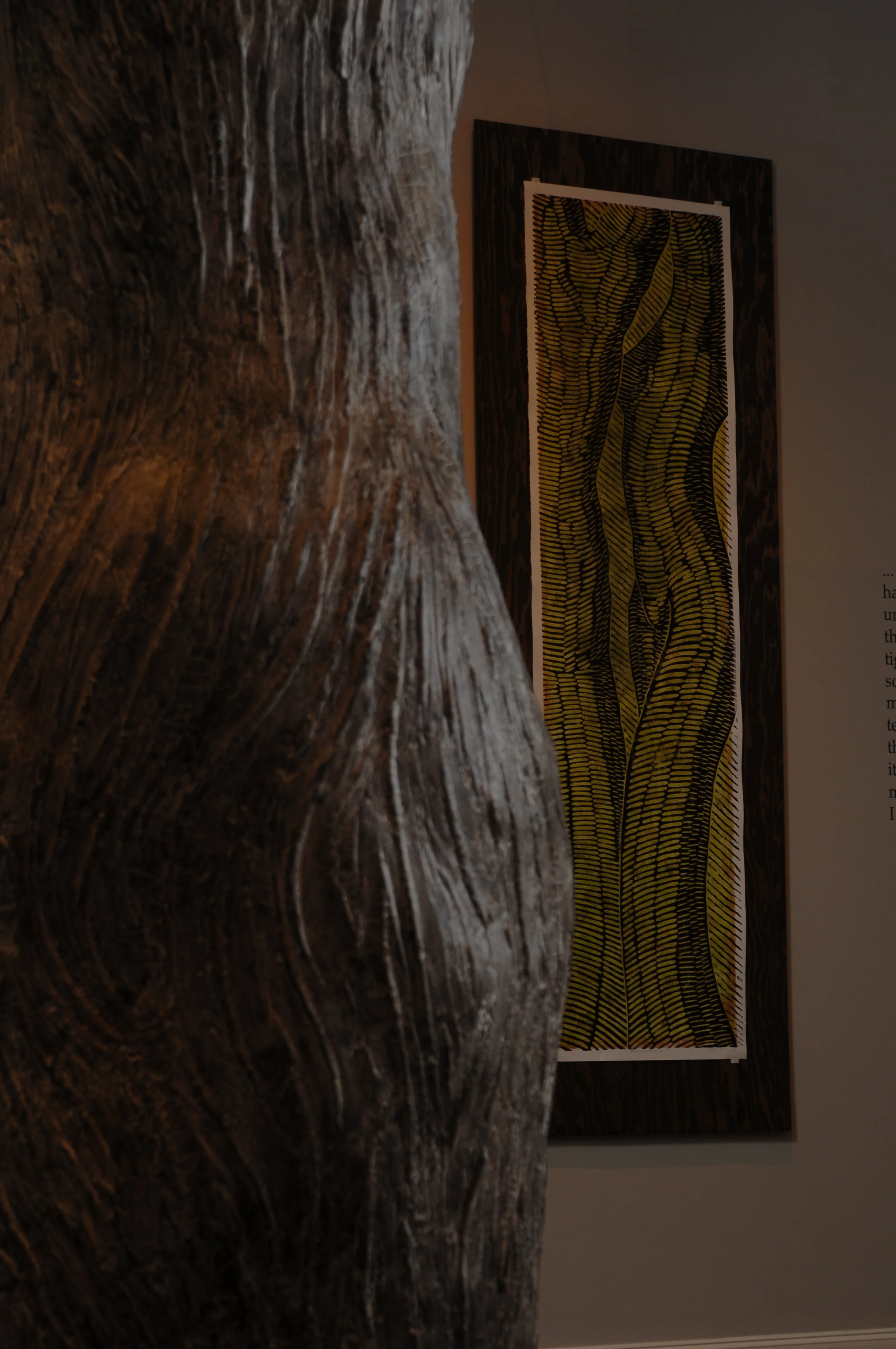 Close-up view of a textured wooden sculpture in front of a patterned artwork hanging on a wall.