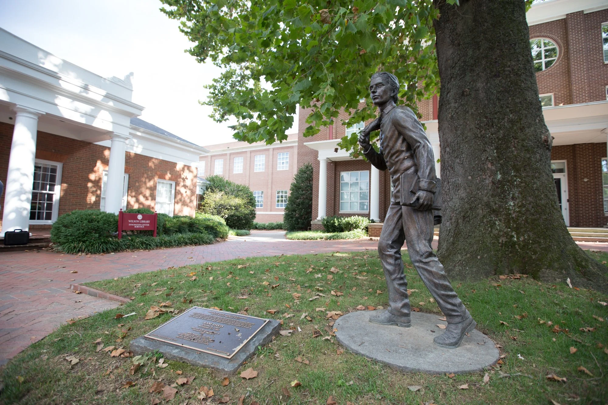 Statue of a man holding a book, located under a large tree outside a brick building with white columns and windows. There is a plaque on the ground in front of the statue and a purple sign to the left indicating it is part of Wilson Library.