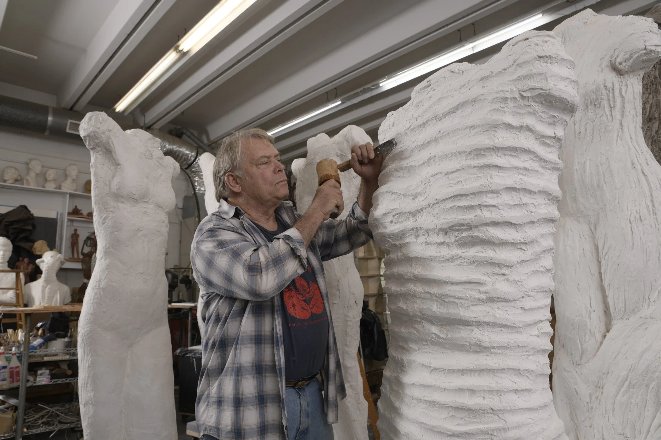 An older man in a plaid shirt carving large white sculpture with a hammer and chisel in an art studio. The sculpture has wavy designs and he is focused on his work.
