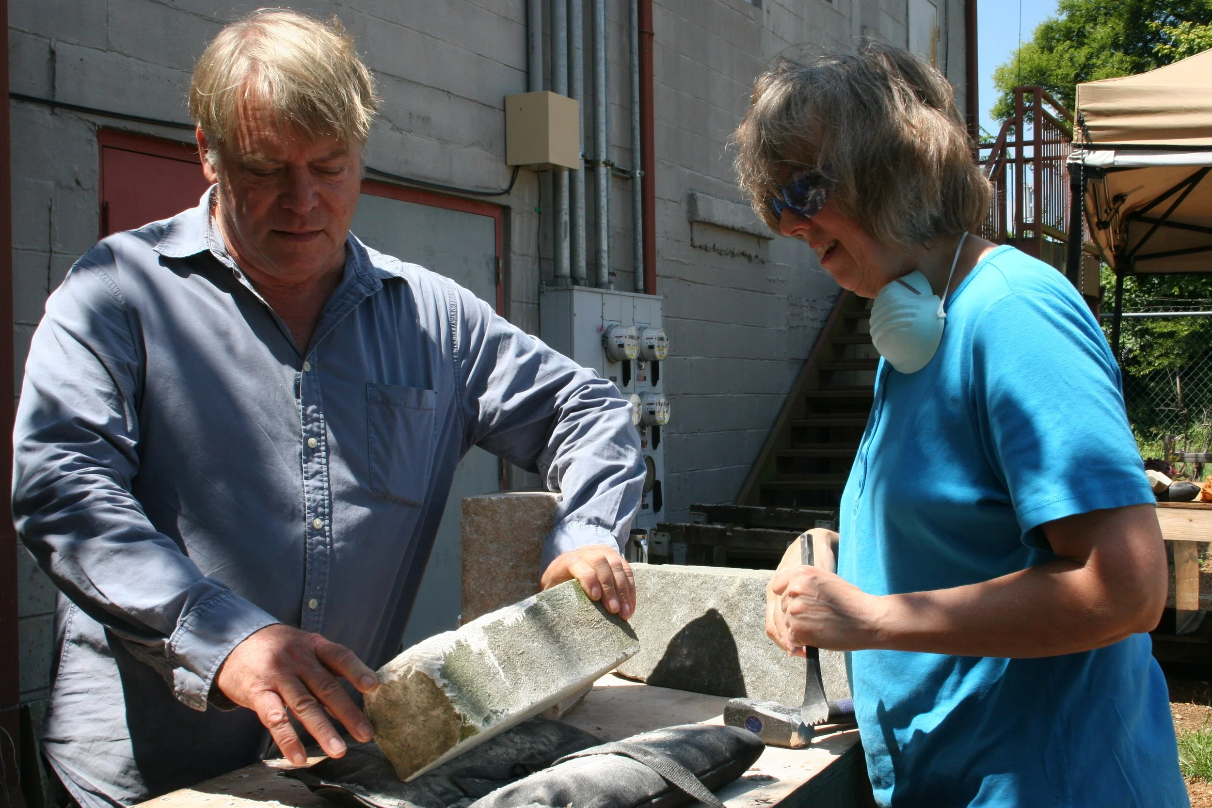 Two people working outdoors with bricks. The man on the left is holding a brick, and the woman on the right is smiling while working on a project. They are next to a wall with electrical meters and pipes, and there is a canopy and stairs in the backg