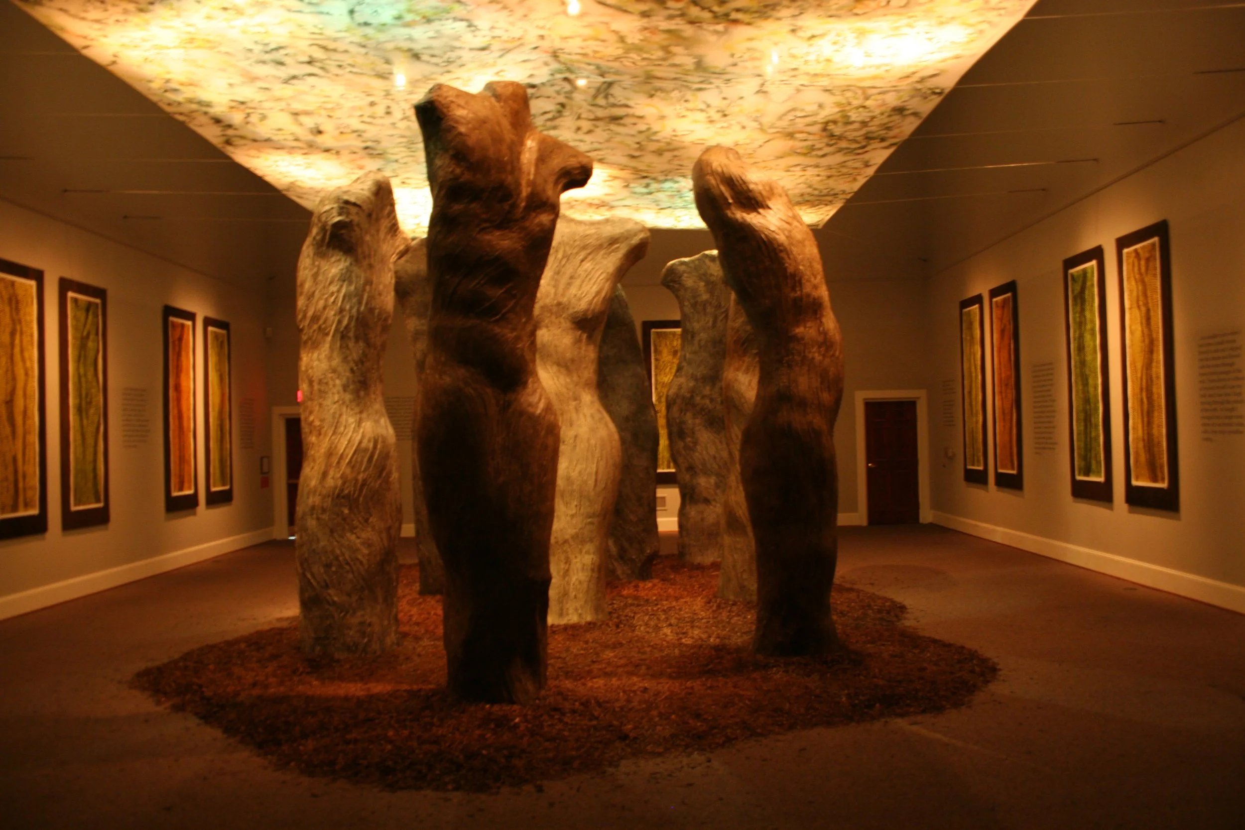 An art installation features several wooden tree-like sculptures arranged in a circle on a carpeted floor, with framed artwork hanging on the walls and a cloud-like ceiling projection. Dream Forest Installation. 