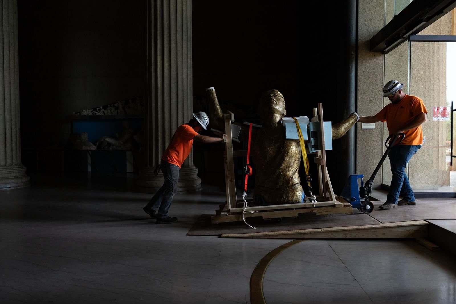 Two workers in orange shirts and white helmets move a large, gold-colored sculpture of a human head and torso on a wooden platform with straps, inside a dimly lit building near an open door leading to outside.