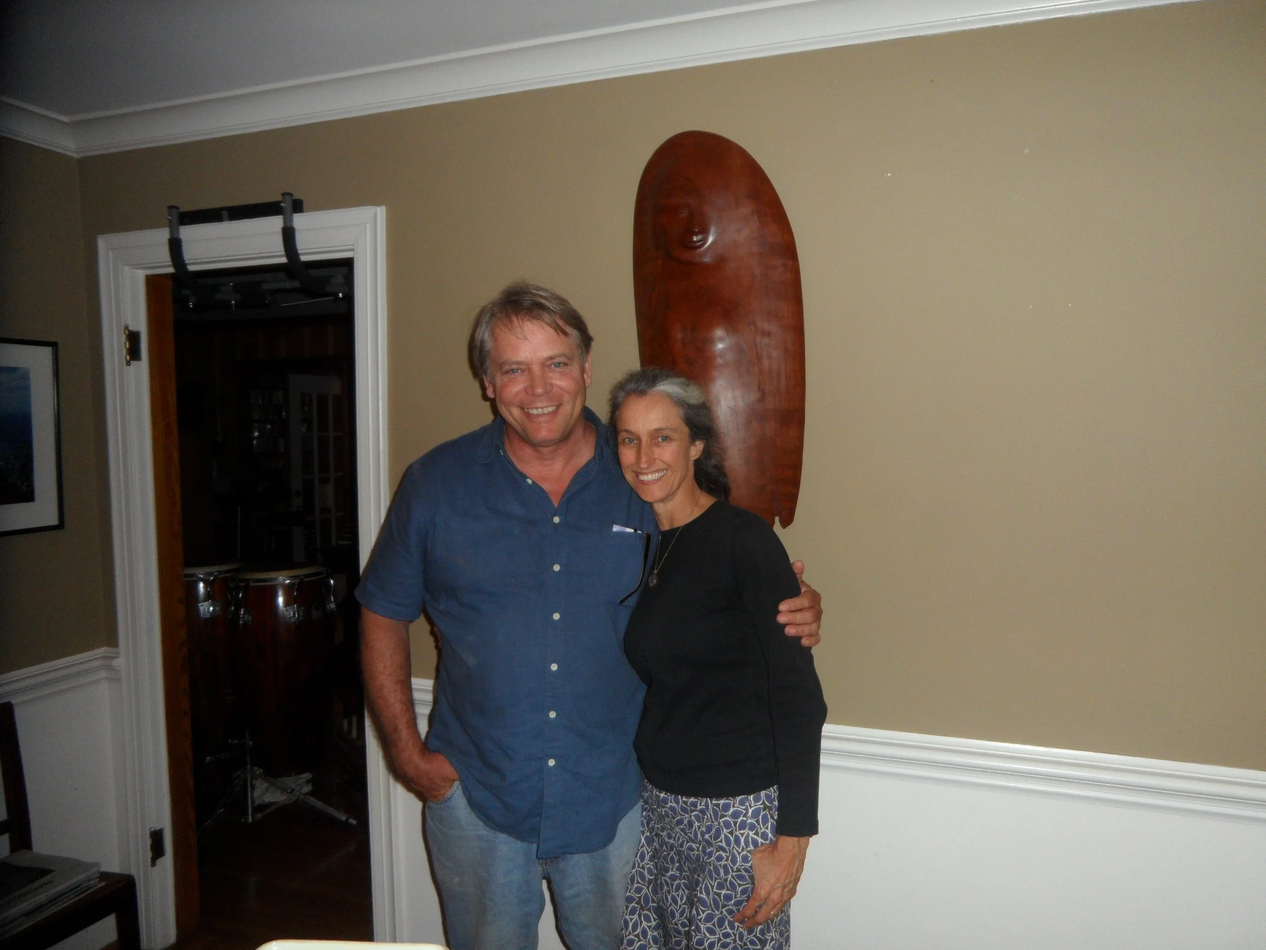 Two smiling adults, a man and a woman, stand together in a room with beige walls. The man has light brown hair and is wearing a blue shirt. The woman has gray hair and is wearing a black top with a patterned skirt. Behind them, a wooden African mask with a face carved into it hangs on the wall.