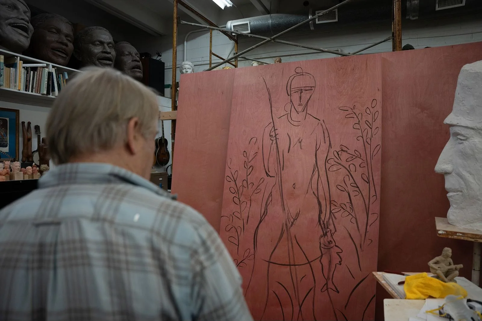 An older man with gray hair wearing a plaid shirt observes a large canvas with a line drawing of a person holding a fish, surrounded by plants, in an artist's studio filled with sculptures, busts, and masks.