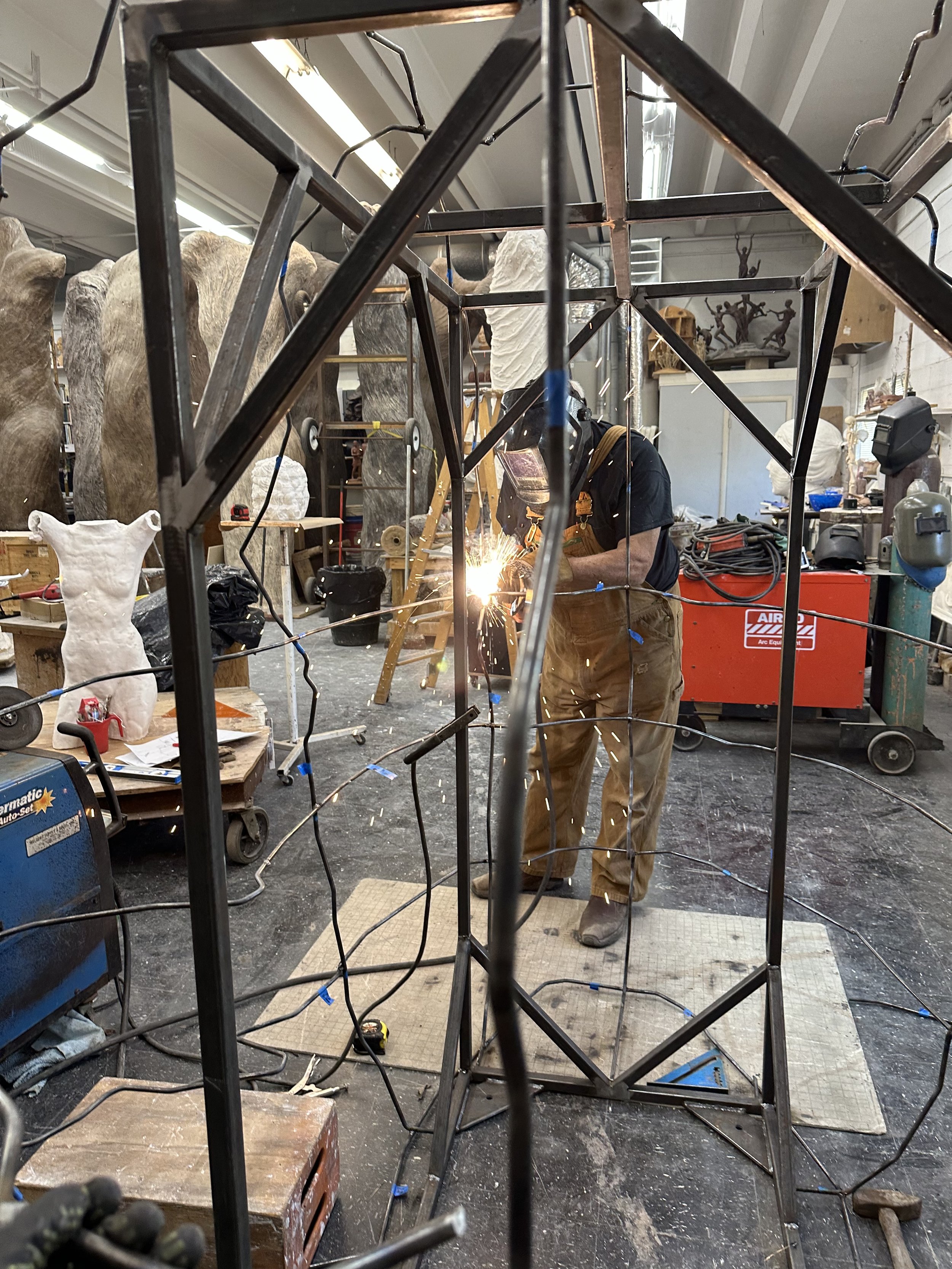 A worker welding a metal structure surrounded by various tools and materials in an art studio or workshop, with sculptures and art pieces visible in the background.