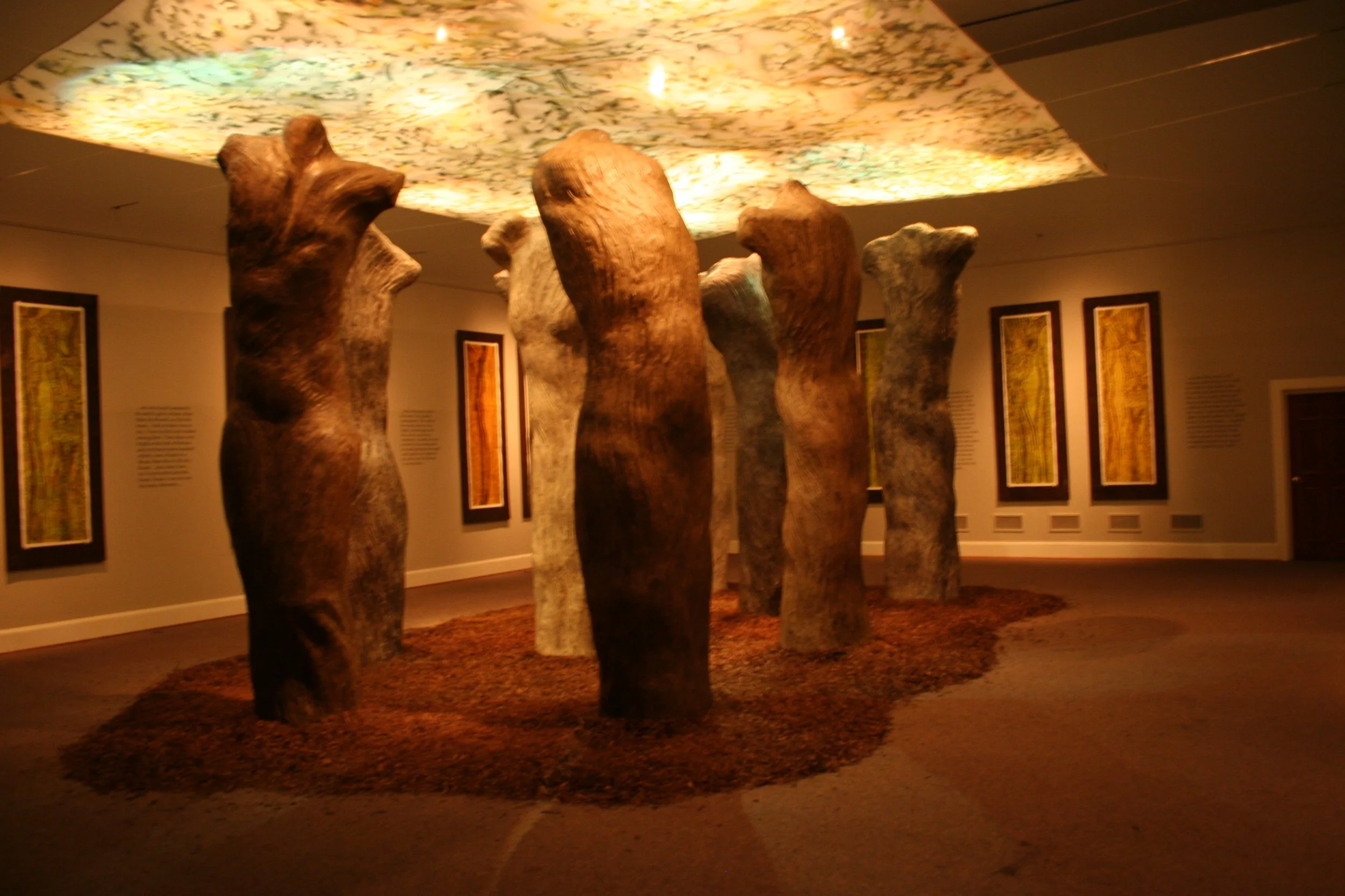 A museum display of tall, carved wooden sculptures resembling human figures standing on a reddish-brown floor with a textured sculpture on the ceiling above. Dream Forest Installation.