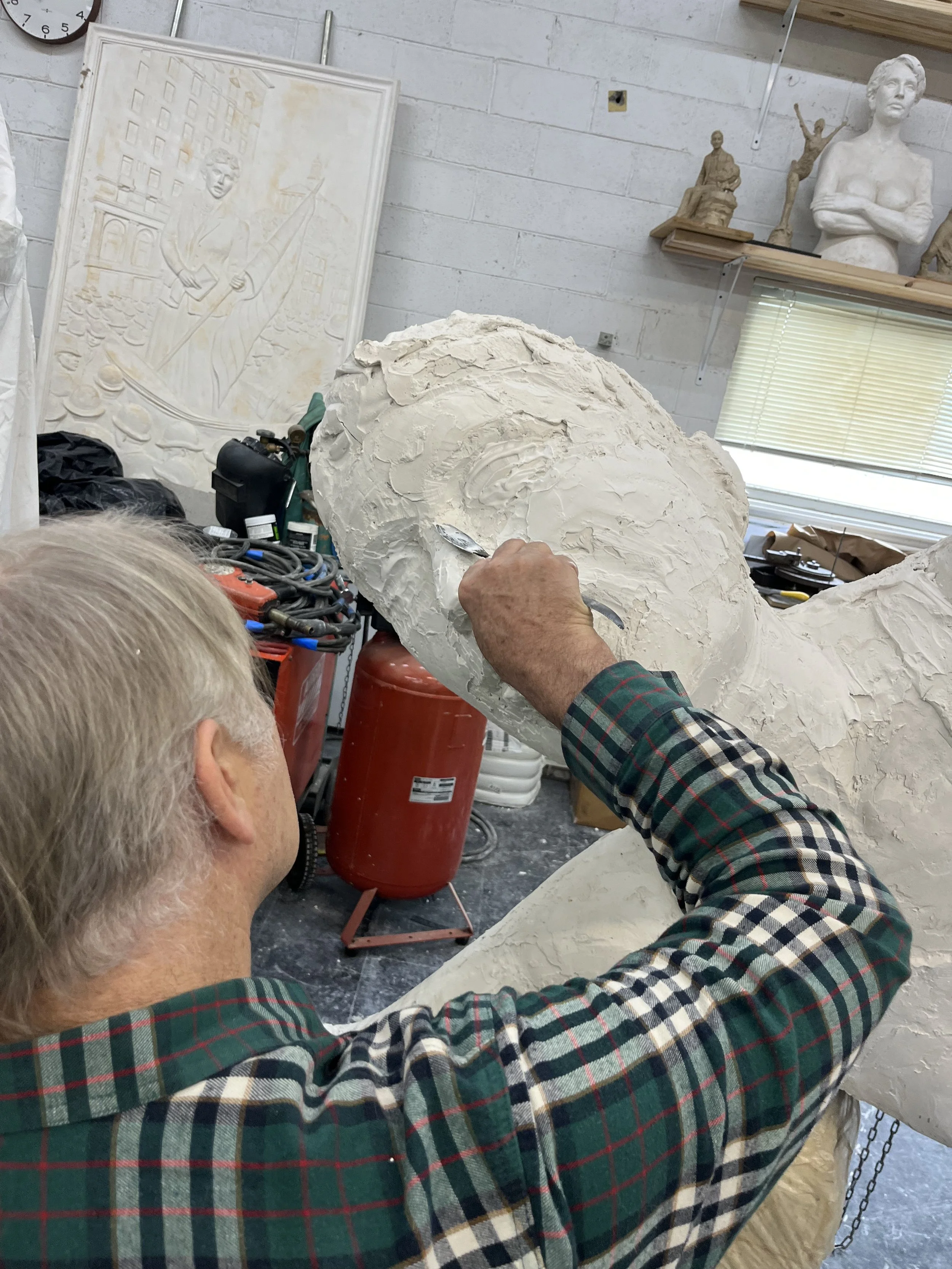 A person working on a sculpture in a workshop, focusing on the head of a large sculpture or bust, with tools and equipment visible, and various art sculptures and artworks in the background.