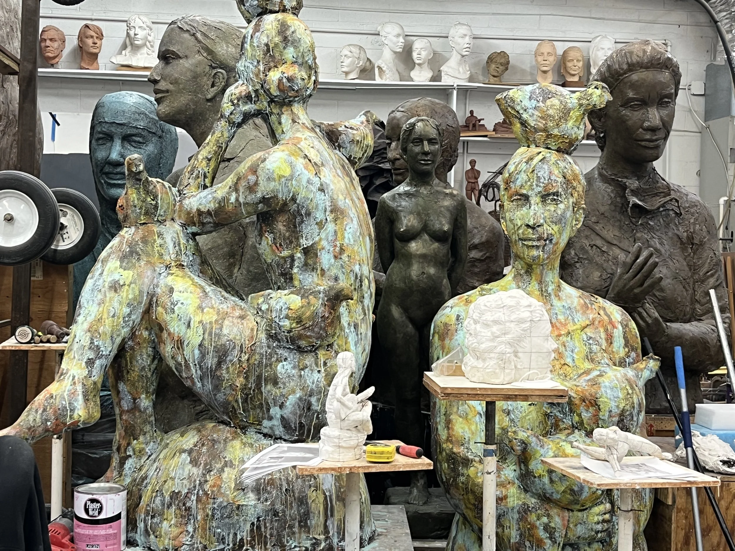 A workshop with multiple sculptures of human figures in various stages of completion, including busts and full-body statues, with some painted in colorful abstract styles. Shelves behind hold busts and small sculptures, and tools and supplies are vis