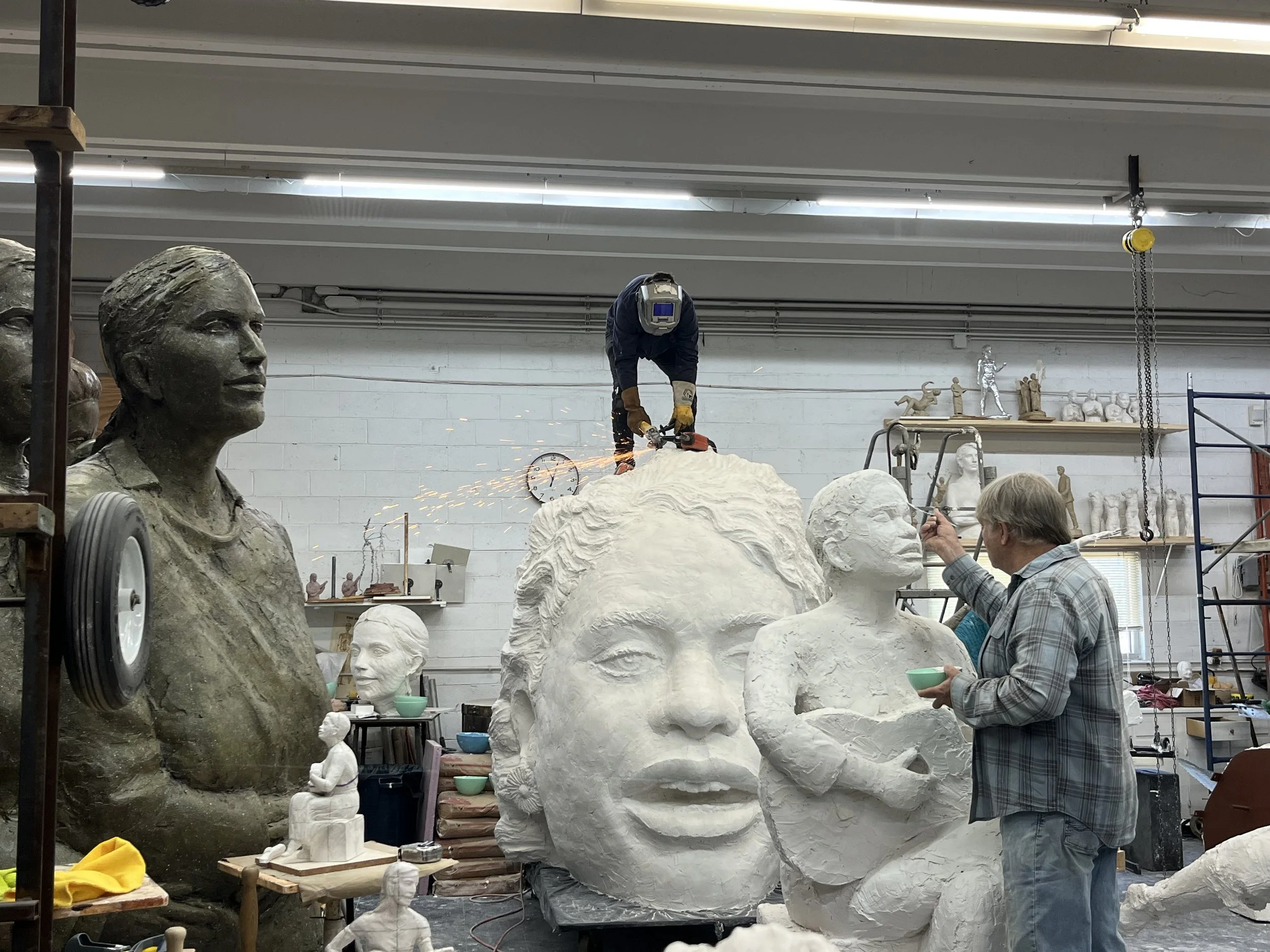 An artist workshop with sculptures of human heads, busts, and figures. A person in safety gear is working on a sculpture while an older man is observing and holding a bowl.