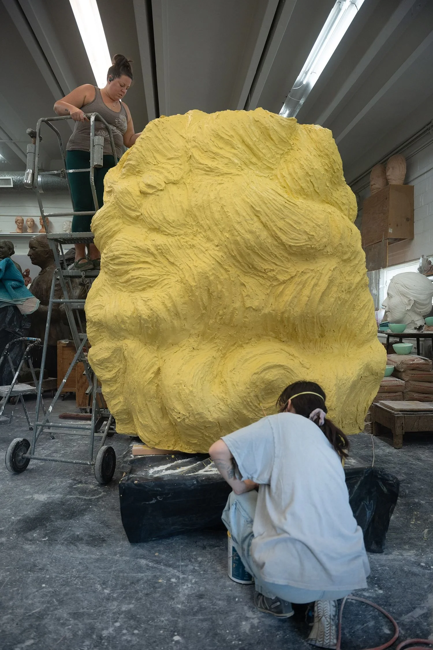 A woman standing on a stepladder working on a large yellow sculpture with a textured surface in a studio, with a young girl crouching nearby.