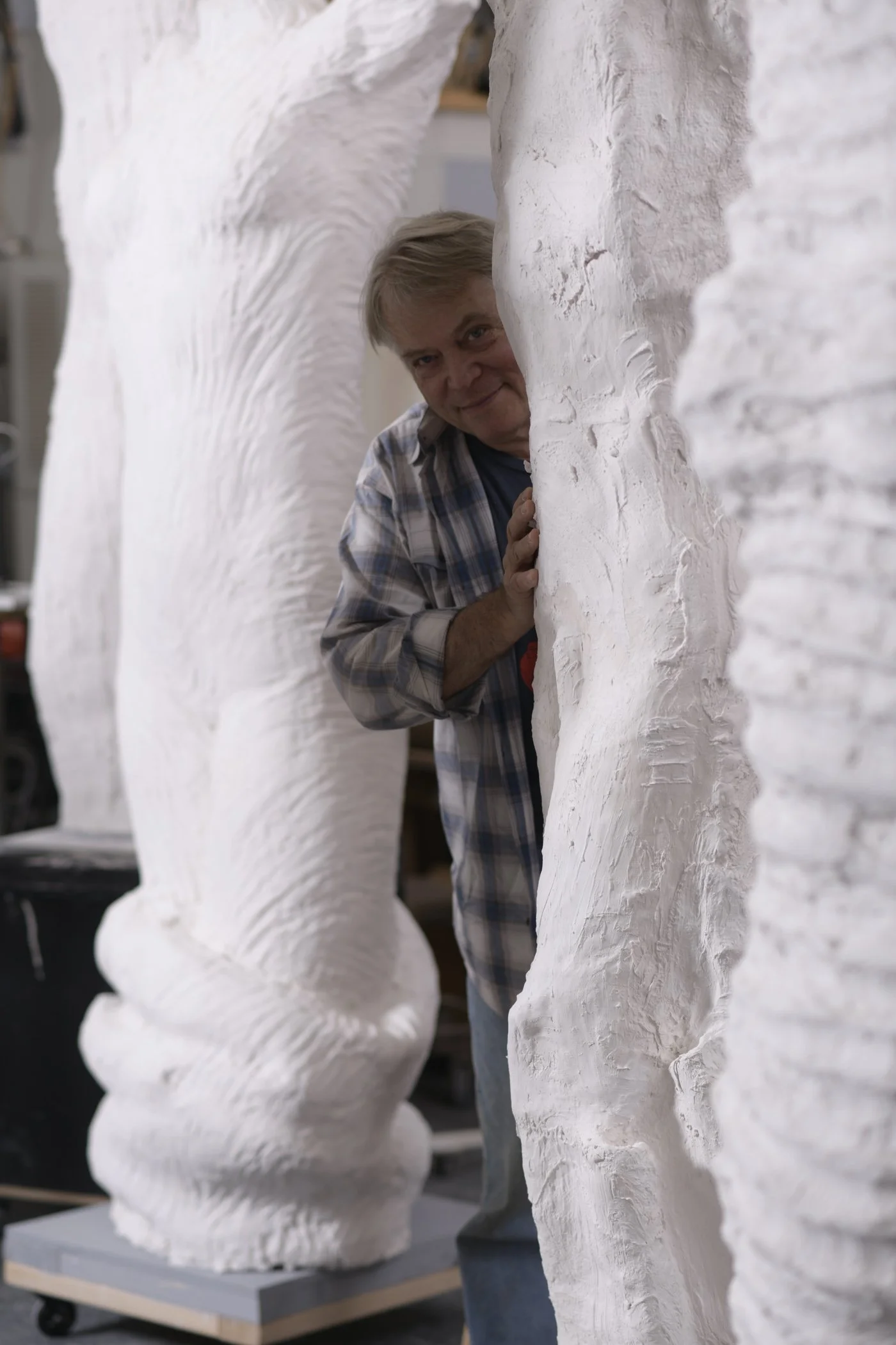 A man peeking between two large white sculptural forms in an artist's studio.