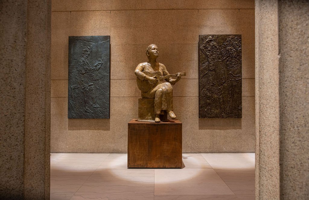 A sculpture of a seated woman playing a guitar, displayed on a wooden pedestal in an art gallery with two bronze relief panels on the wall behind her.