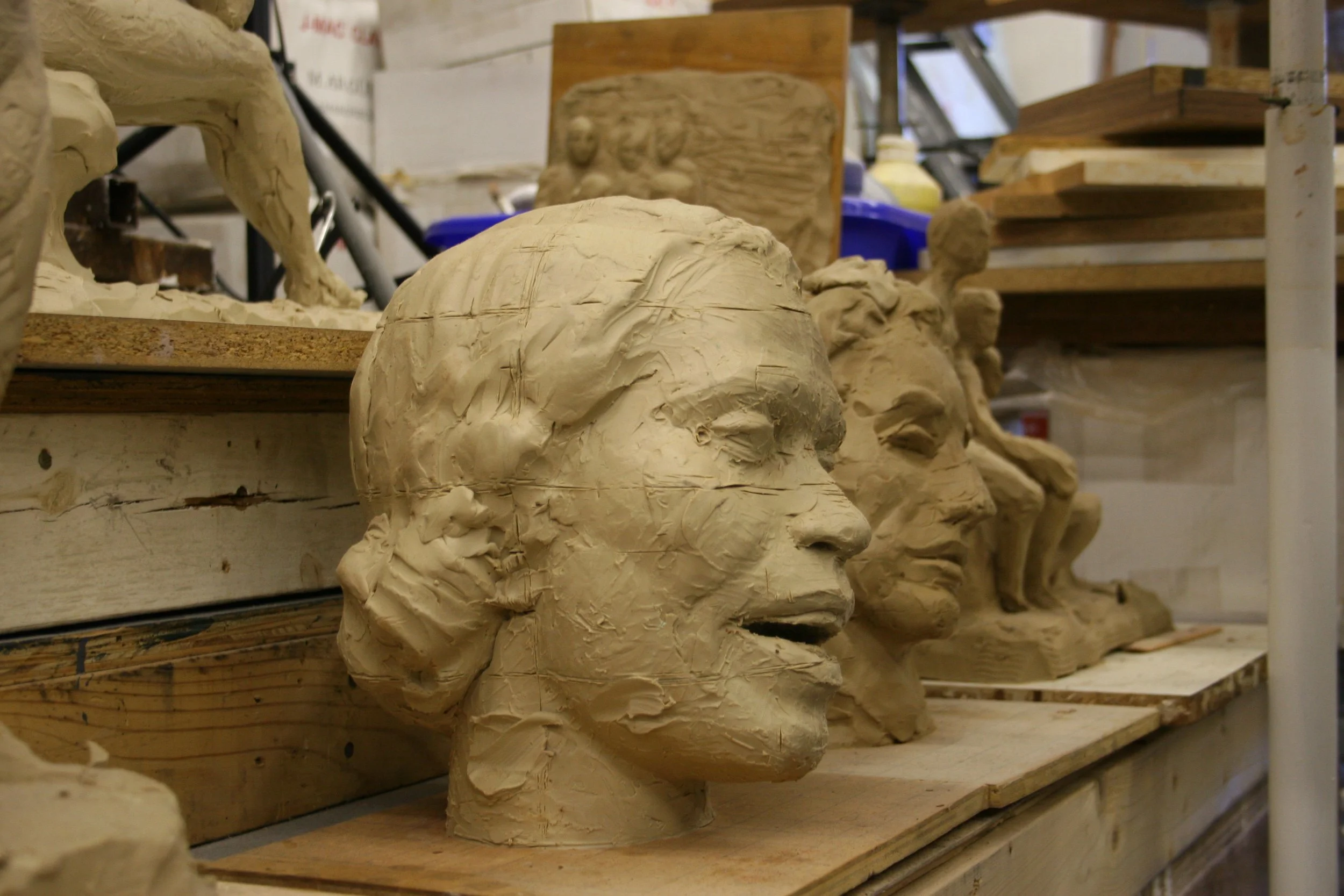 Clay sculpture heads on a wooden workbench in an art studio, with shelves and art supplies in the background.