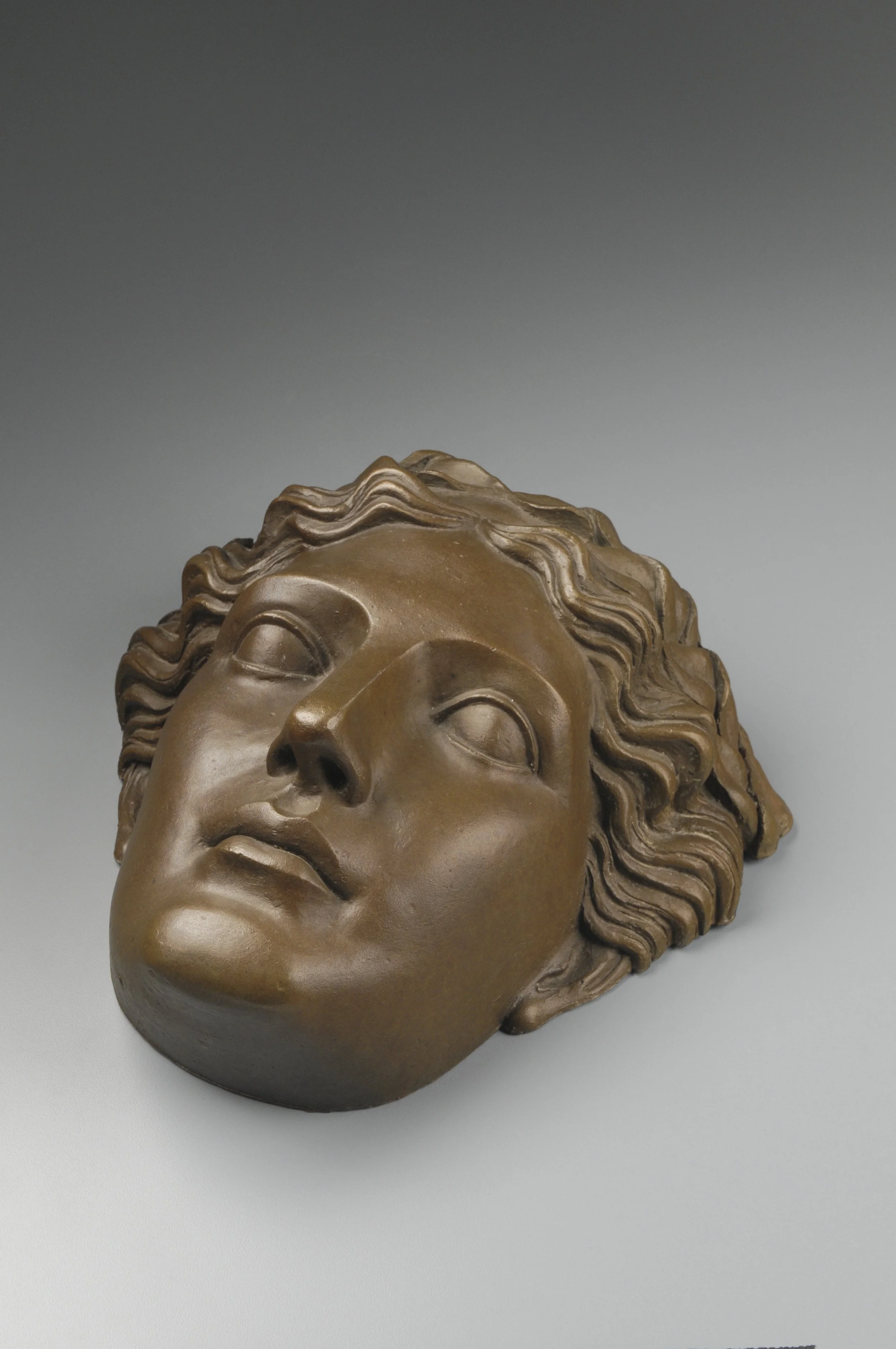 Close-up of a bronze bust sculpture of a woman's face with wavy hair, set against a plain gray background.