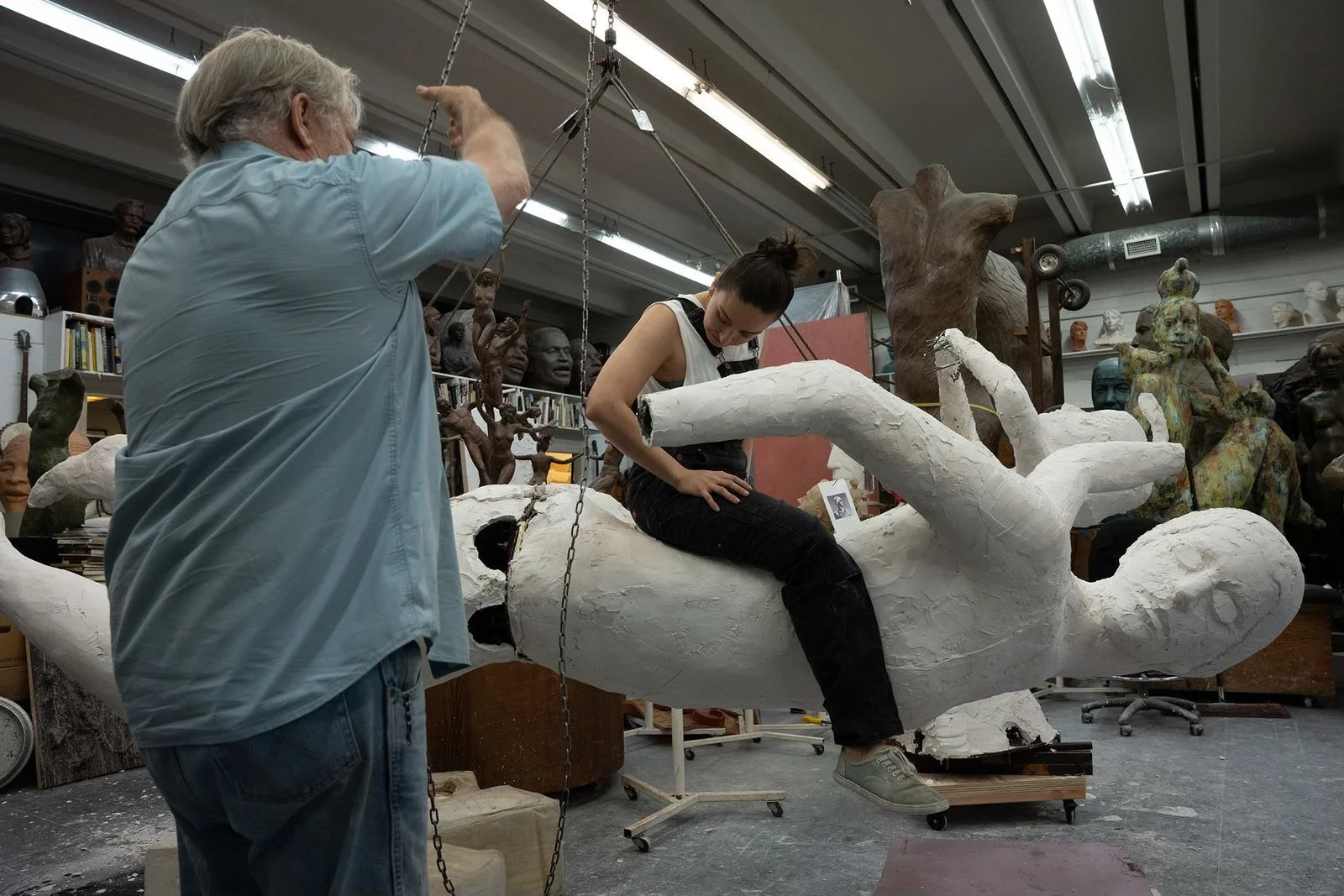 Alan Lequire and Amber Lelli working on a large sculpture of a human figure in an art studio filled with various busts and sculptures in the background.