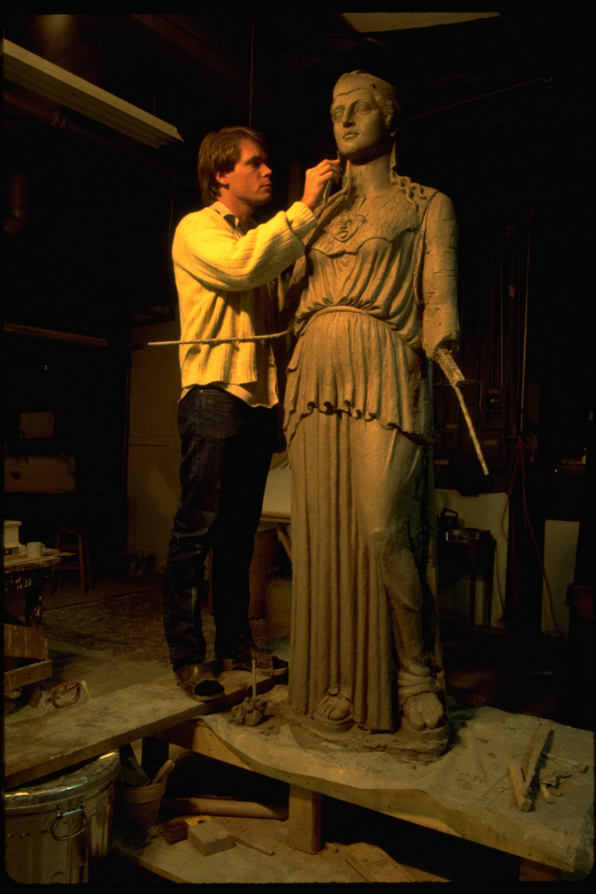A man in a yellow sweater working on a large stone sculpture of a woman, adjusting part of the sculpture's face. Athena Parthenos in Parthenon Museum Nashville. 