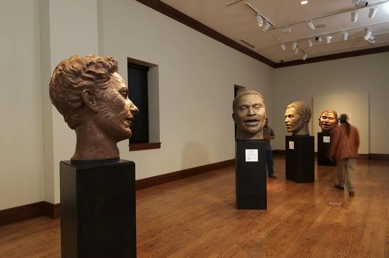 Bronze bust sculptures of famous figures displayed on black pedestals in an art gallery with visitors observing.  Cultural Heroes