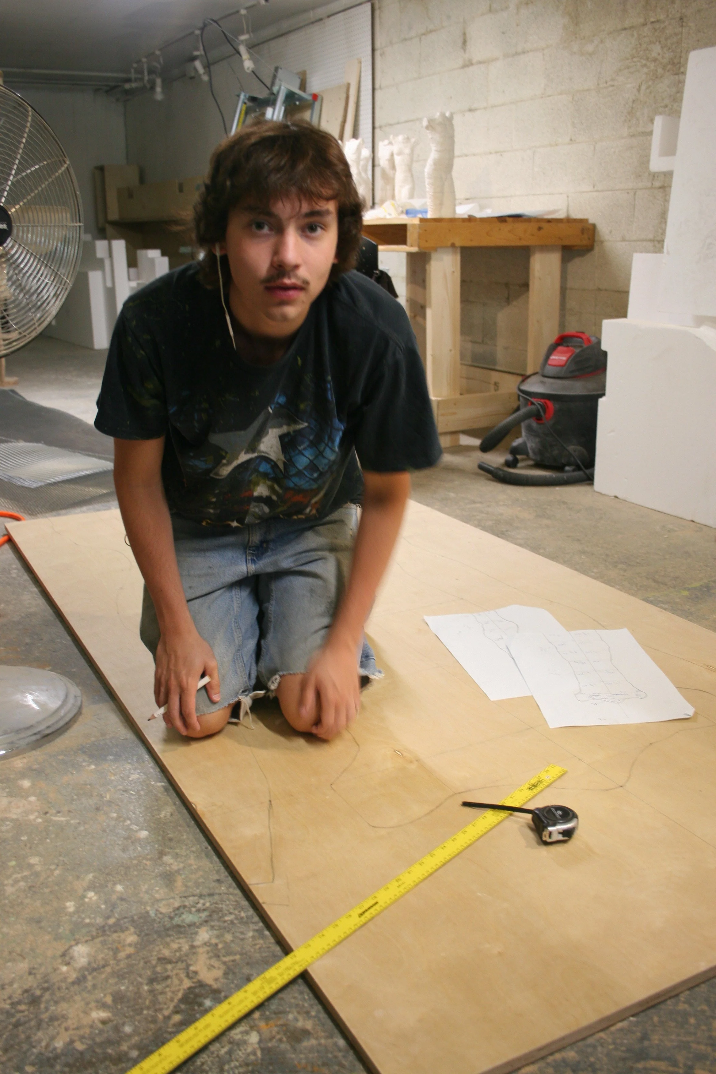 A young man with brown hair and a mustache kneeling on the floor of a workshop, surrounded by tools and materials, with a sheet of paper with a sketch in front of him. He appears to be working on a project, with a measuring tape and a ruler nearby.