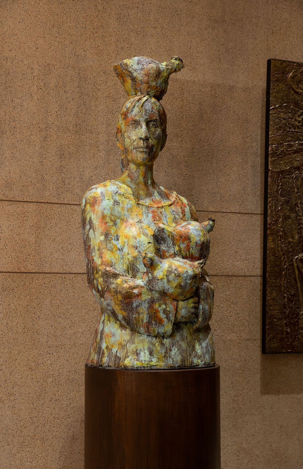A colorful sculpture of a woman holding two chickens, with a bird perched on her head, displayed on a pedestal against a plain wall.