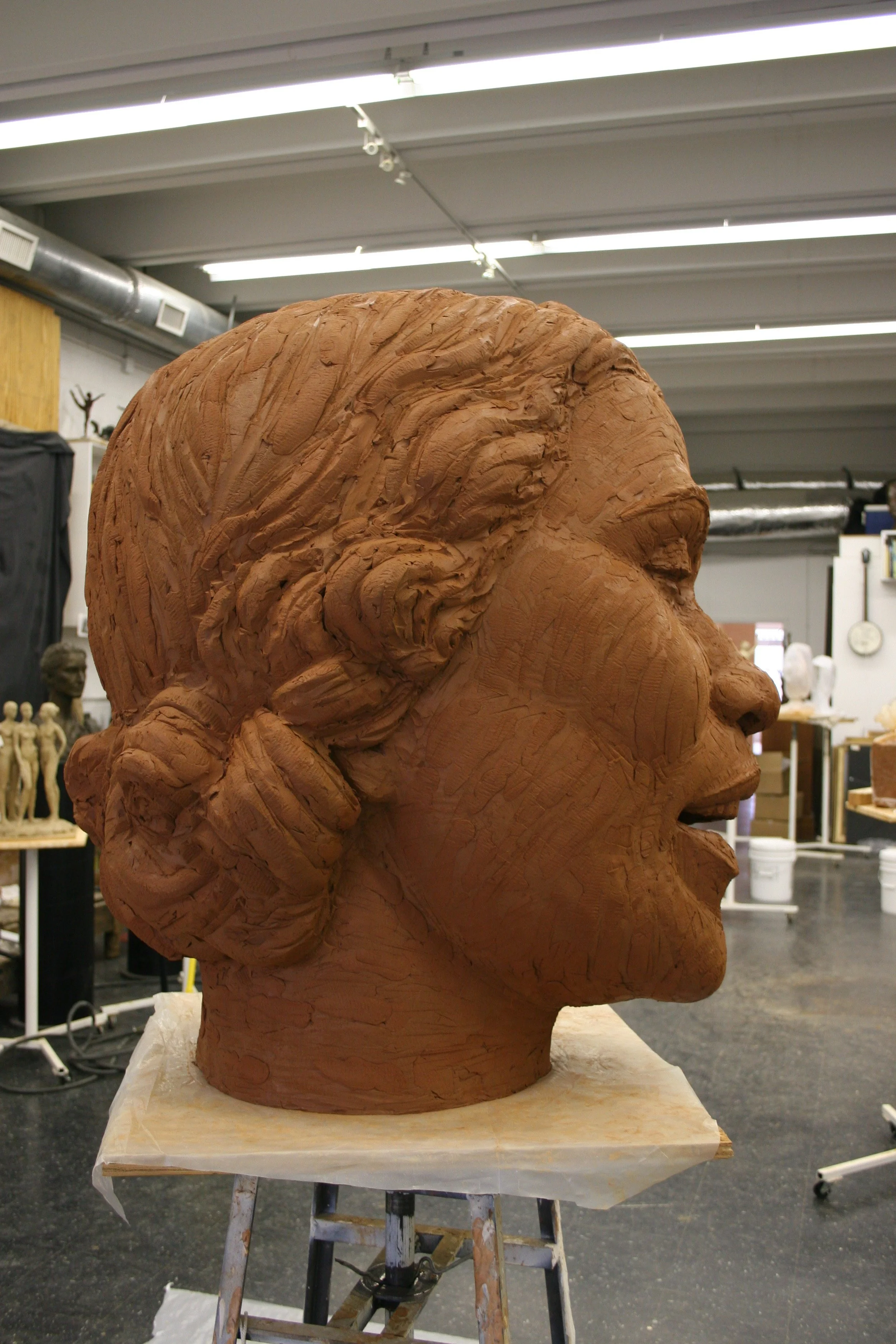 A large bust sculpture of a person's head made of clay is in a studio. The sculpture depicts a woman with hair styled in a bun, and a hand resting on her temple. Shelves with smaller sculptures and art supplies are visible in the background.