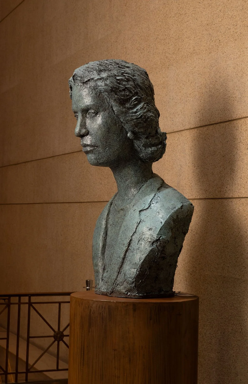 Bronze bust sculpture of a woman displayed on a wooden metal against a beige wall.