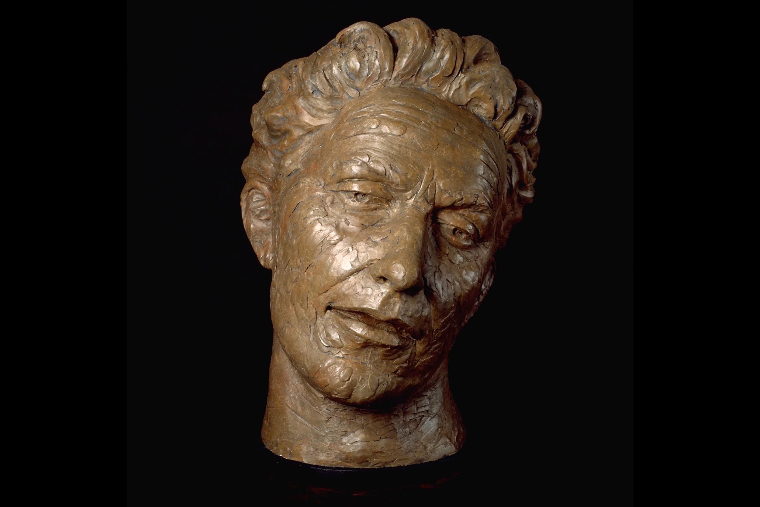 Bronze sculpture of a woman's head with closed eyes and styled hair, set against a black background.