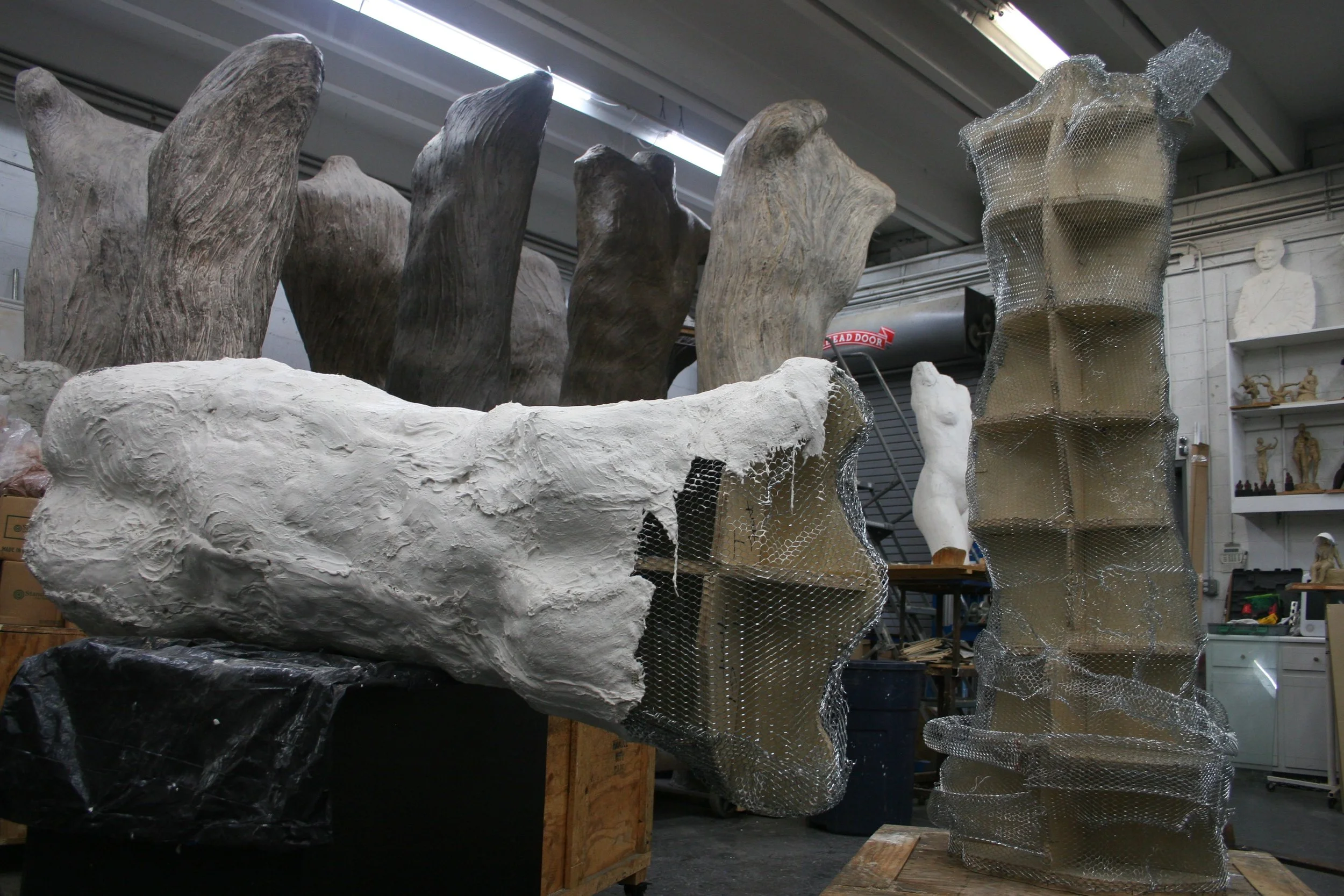 Sculptural art pieces in a workshop, including a piece resembling a hand with fractured forms, some wrapped in metallic wire mesh, and wooden crates and sculptures in the background.