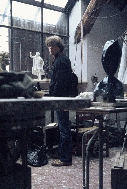 A young man standing in a workshop or studio with sculptures and art tools on the tables.
