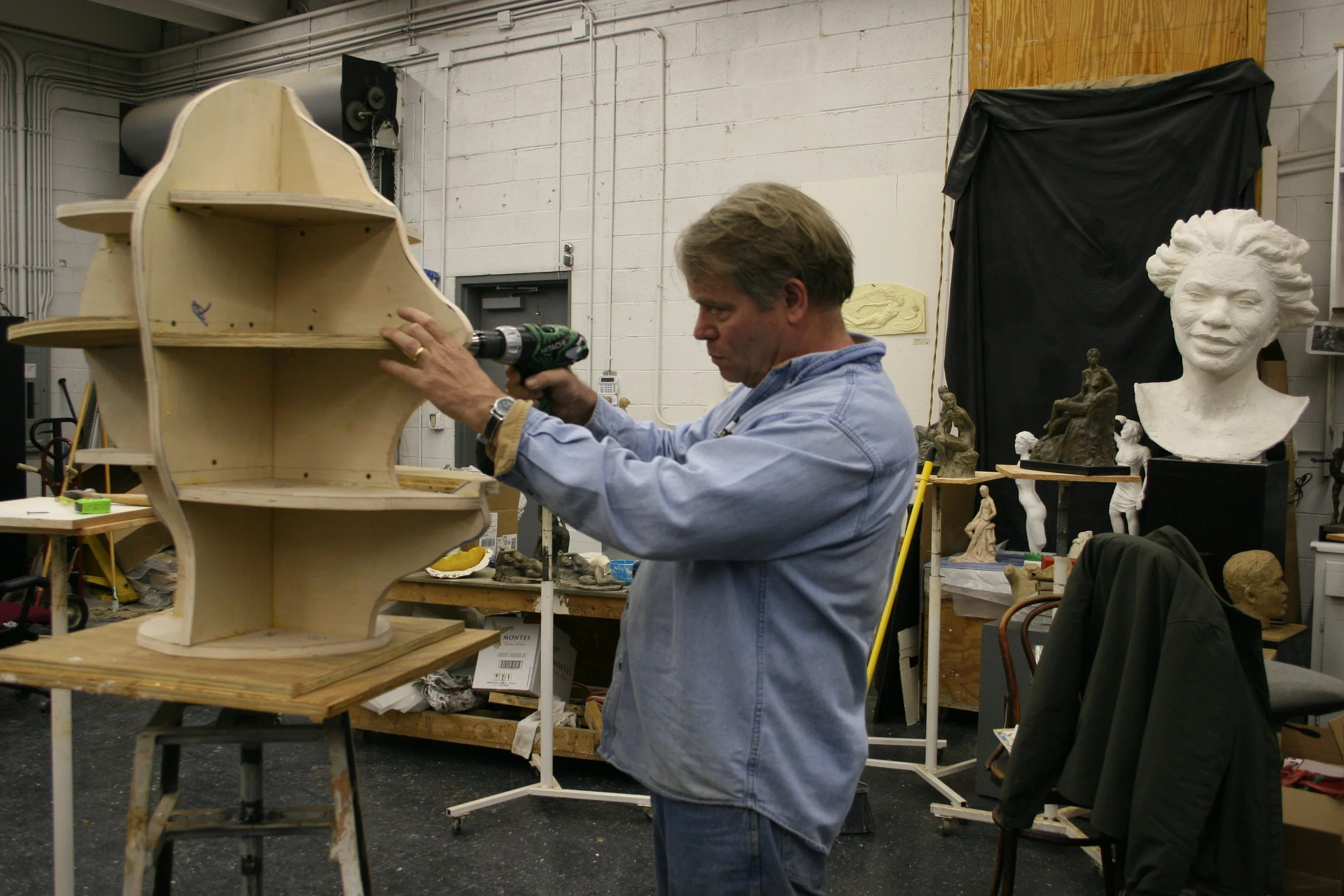 A man Alan Lequire working on a wooden armature sculpture in an art studio surrounded by sculptures and art supplies.