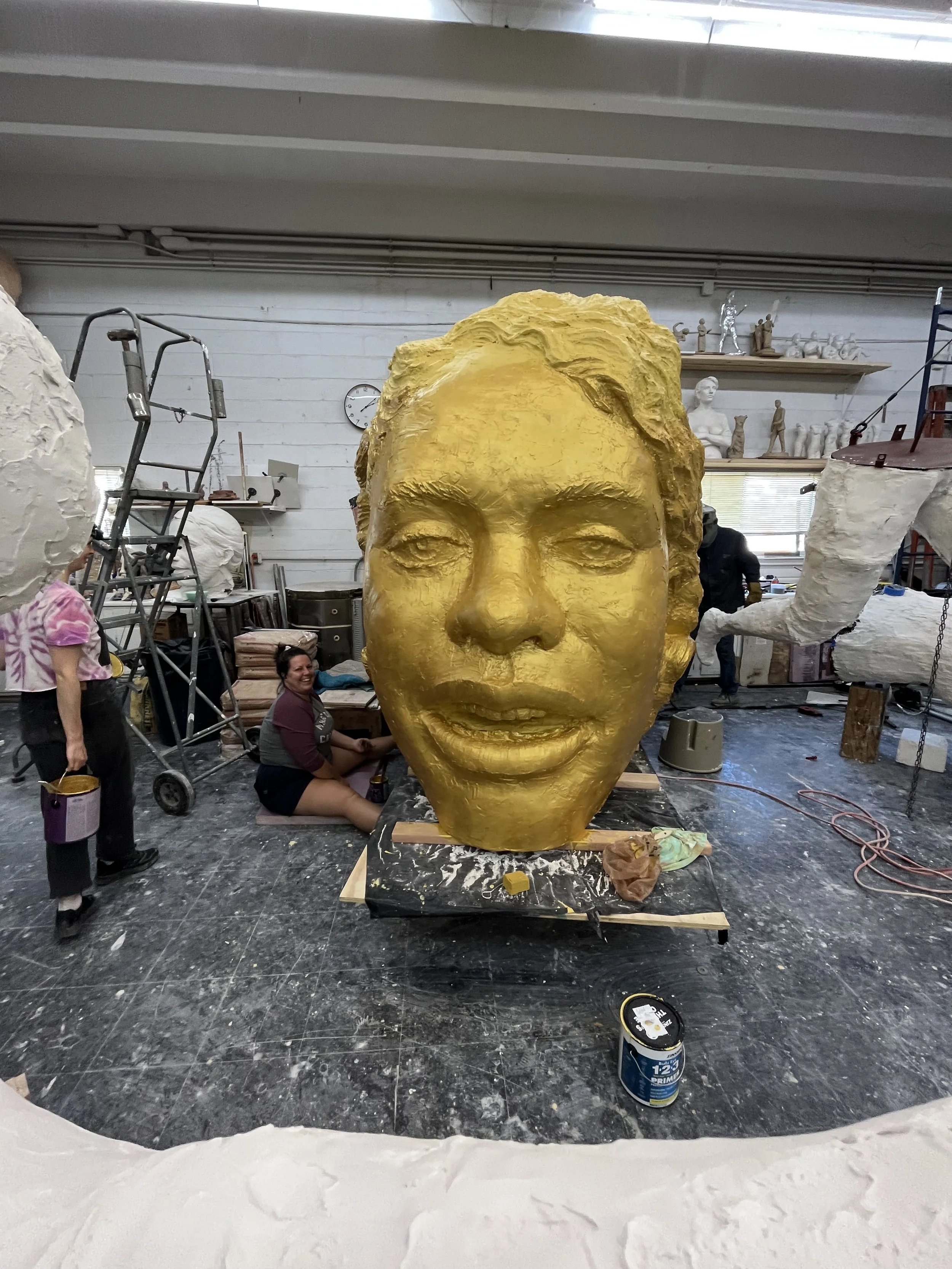 A large artist's sculpture of a human face with a gold finish, set in an art studio with people working and shelves with small sculptures in the background.
