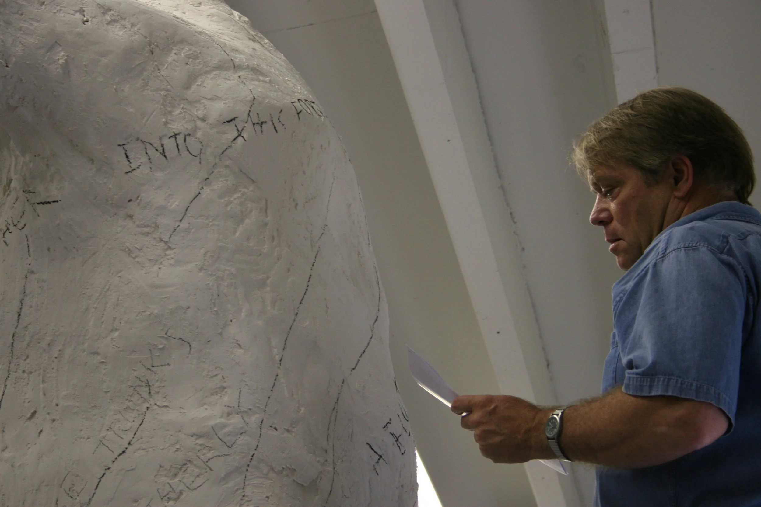 A man with gray hair and a watch on his left wrist, wearing a blue shirt, is writing or carving words on a white, textured, curved wall with black ink or paint. The words are partially visible, with some characters readable, including 'INTO' and 'AN'