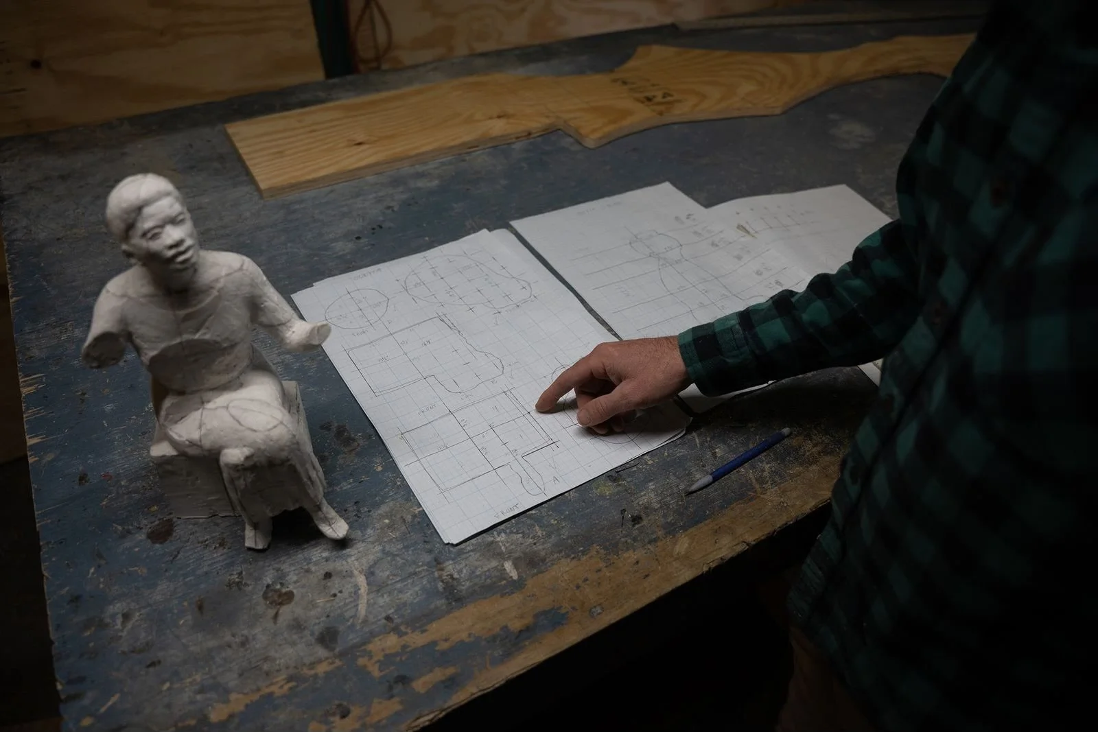 A person in a green plaid shirt looks at a sketchbook with technical drawings on grid paper, while a clay sculpture of a person on a chair sits on the workbench nearby.