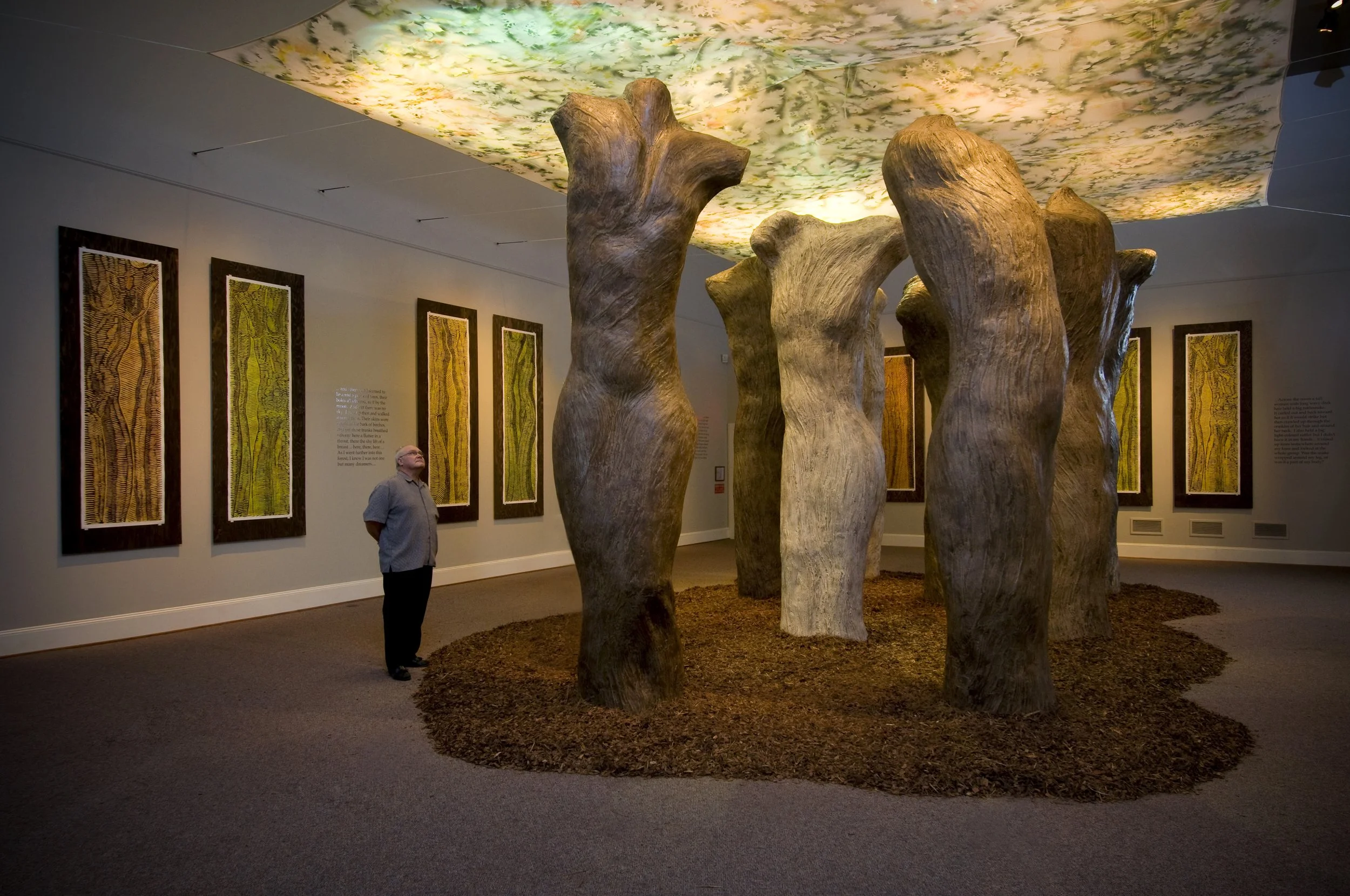 An art installation featuring large wooden sculptures resembling tree trunks in a gallery space. Behind the sculptures are framed artworks on the wall, and the ceiling has a colorful mural. A man is looking at the sculptures.