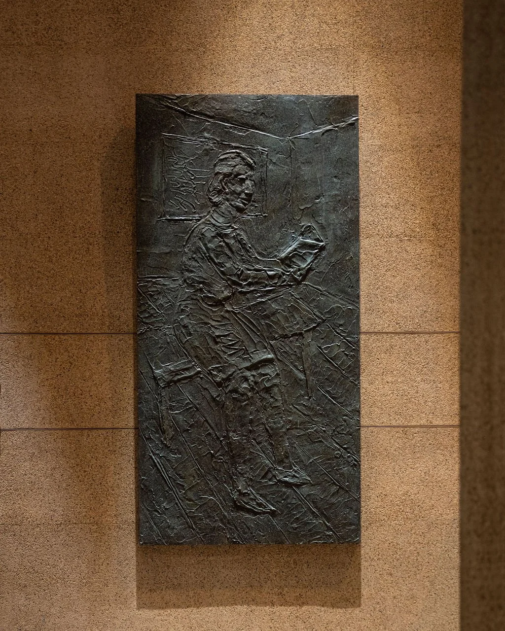 A bronze relief sculpture of a woman sitting and reading a book on a textured background, framed by a wall.