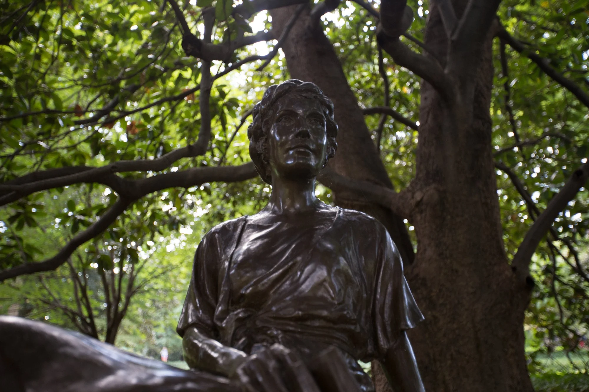 Bronze statue of a woman, outdoors among trees and green foliage.