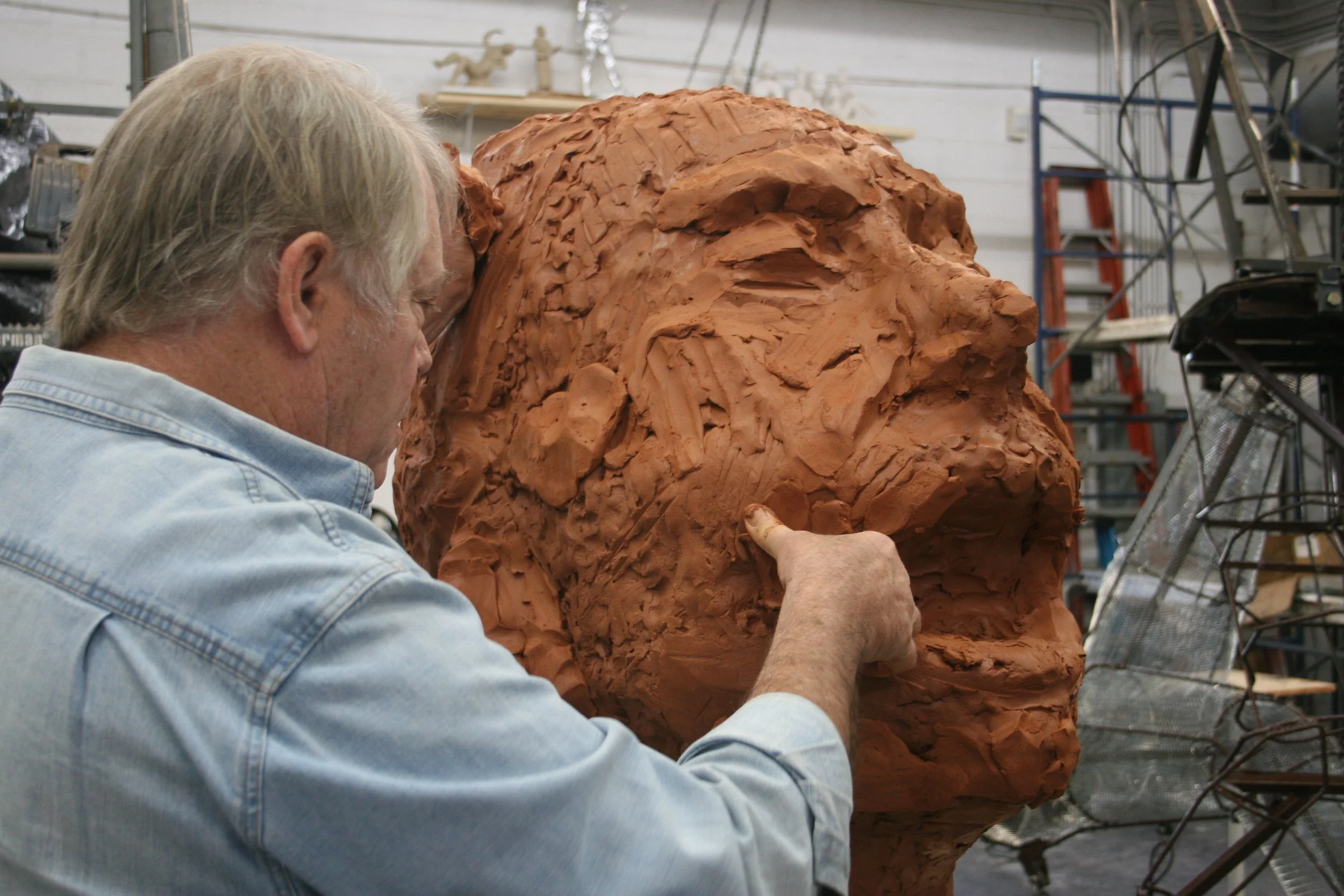 A man with gray hair working on a large clay sculpture of a face in a workshop.