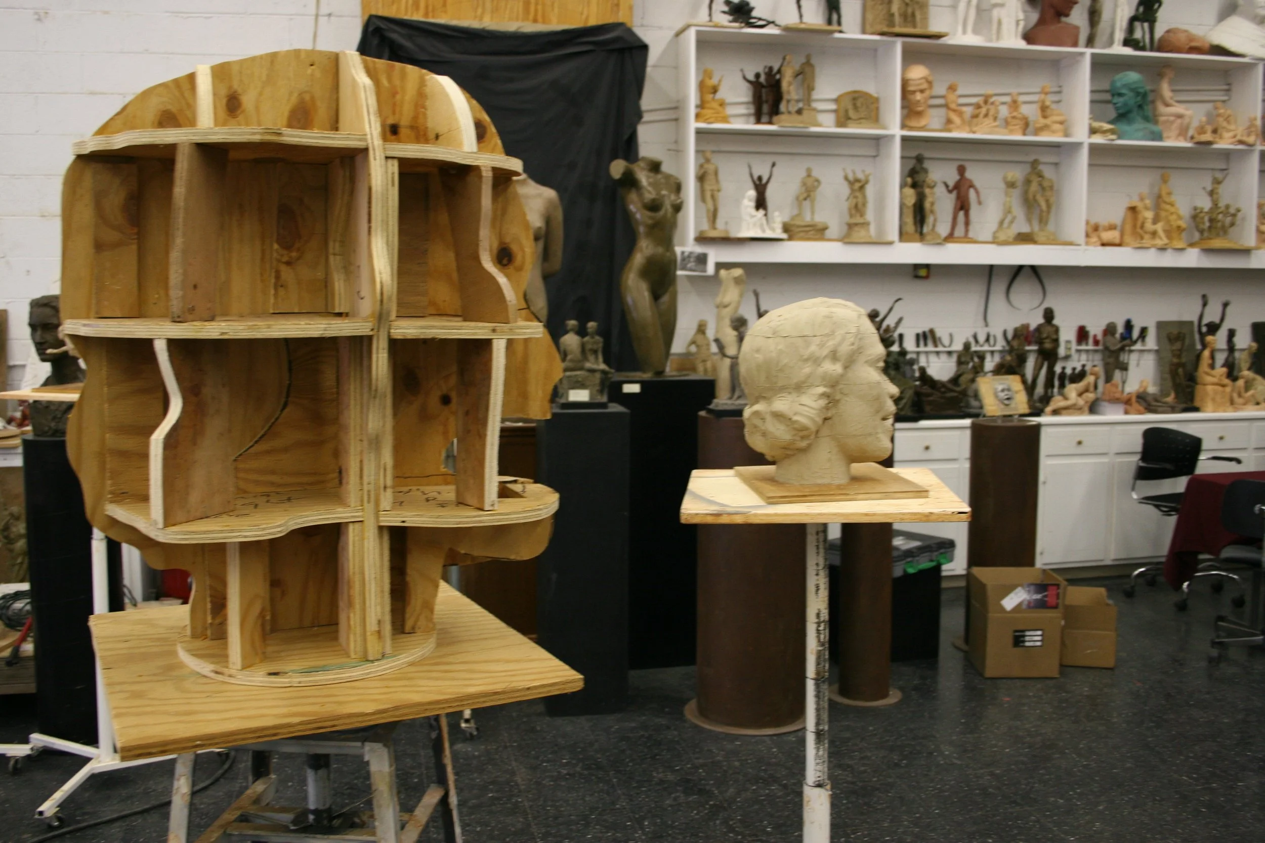 Workshop with various sculptures, busts, and figures displayed on shelves and tables, including a partially built wooden sculpture and a head bust in the foreground.