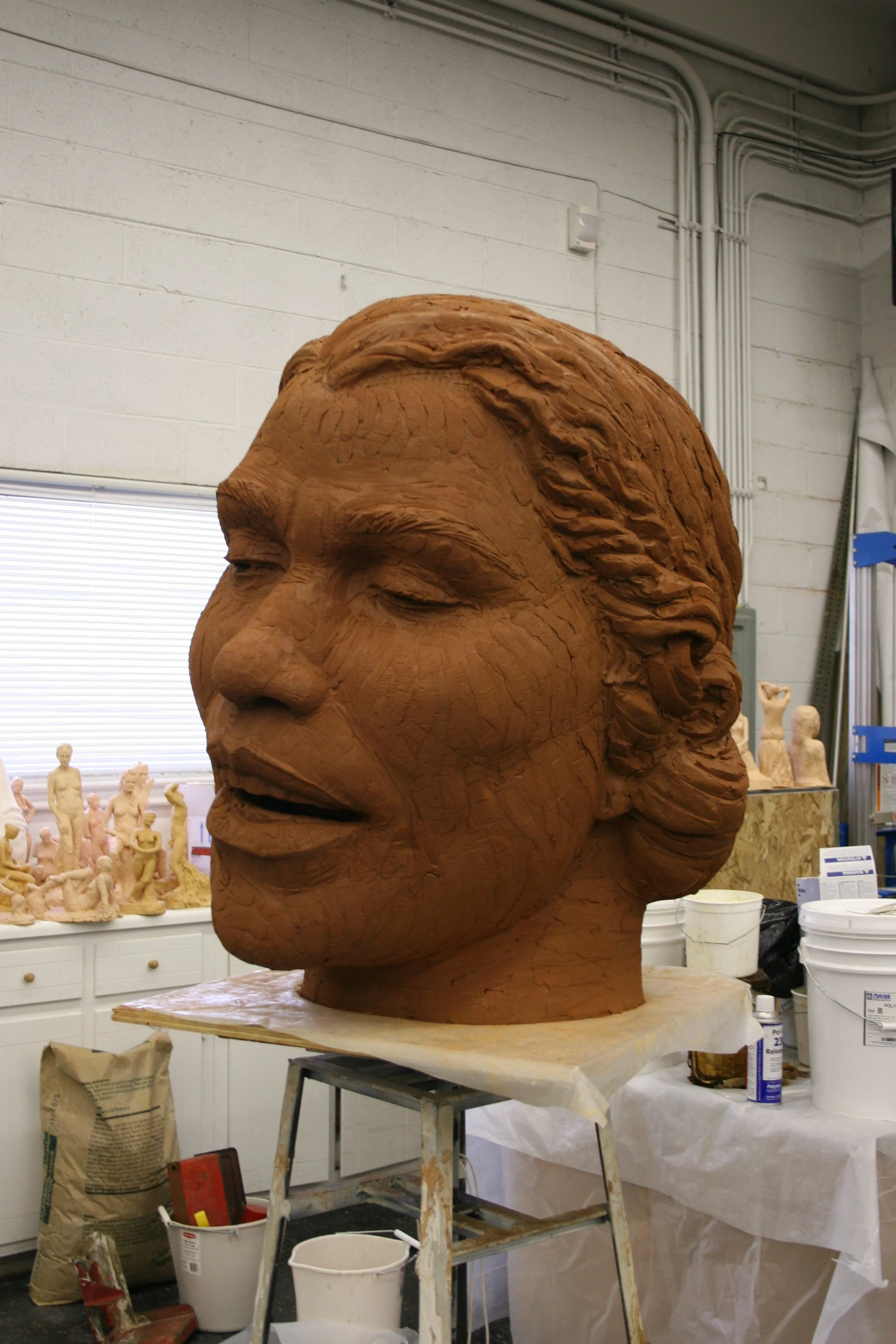 A large wooden sculpture of a person's head is in progress, situated in an art studio with various smaller sculptures and art supplies around.