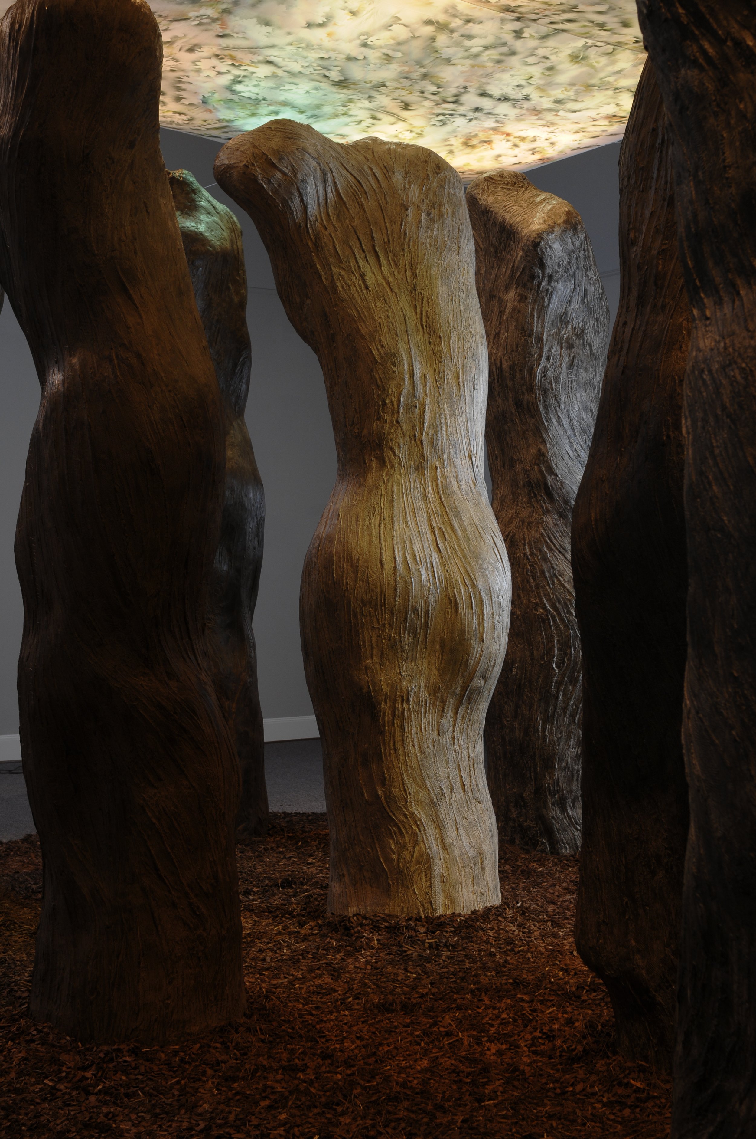 Indoor art installation featuring vertically standing large wooden logs with a textured surface, on a floor covered with wood chips, and an illuminated ceiling resembling a cloudy sky.