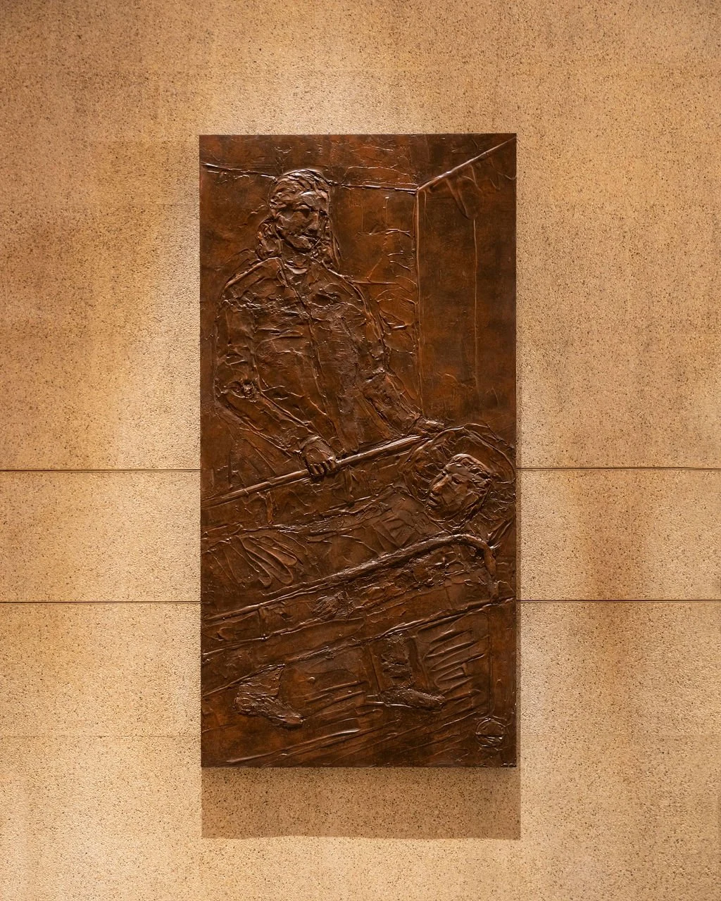 Bronze relief sculpture of female doctor or nurse, one standing with a tool and the other a patient in a hospital bed, mounted on a beige wall.